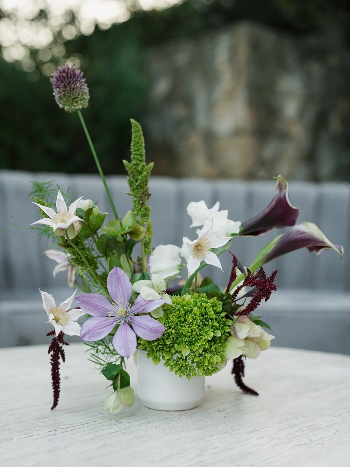  Cocktail hour arrangement of lavender clematis, plum calla lilies, allium and green hydrangea in a white vessel at Camp Lucy 