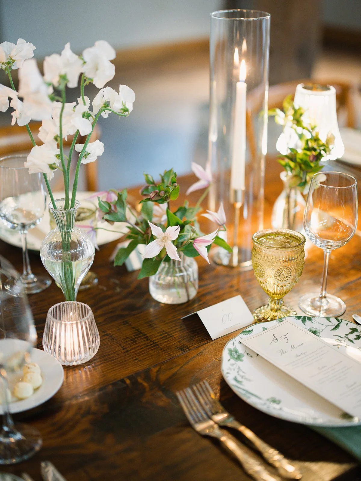  Close-up of white sweet peas, pink clematis and hellebores in bud vases on a candlelit farm table at Camp Lucy in Dripping Springs 