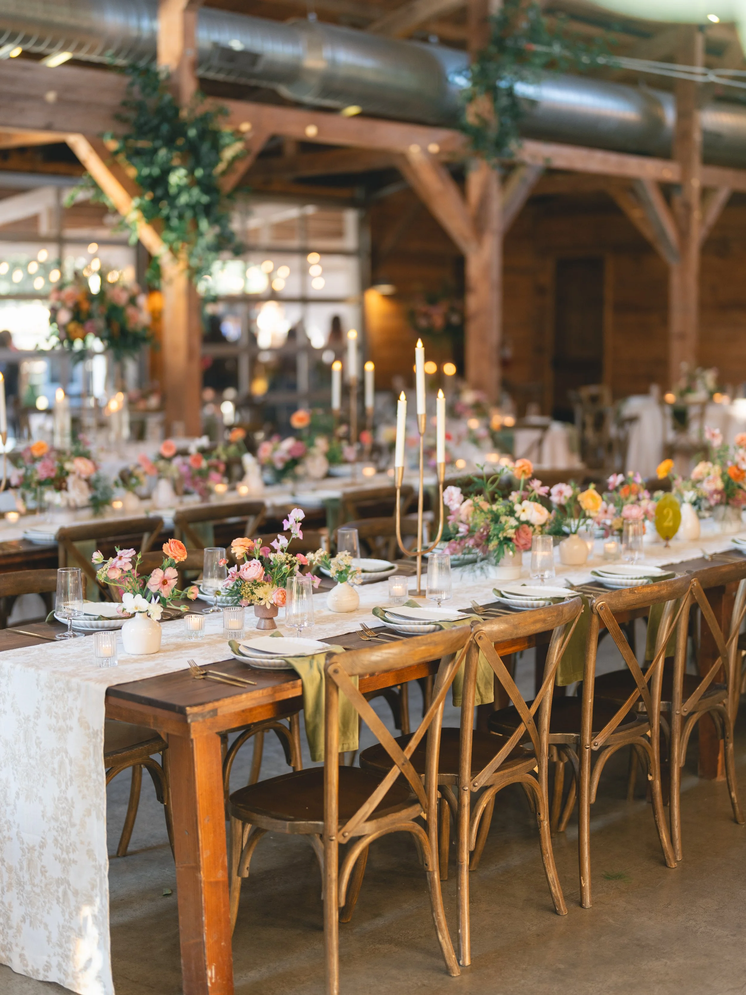  Long farm tables with floral jacquard runners, bud vases of ranunculus and cosmos, and gold candelabras in the barn at The Addison Grove 