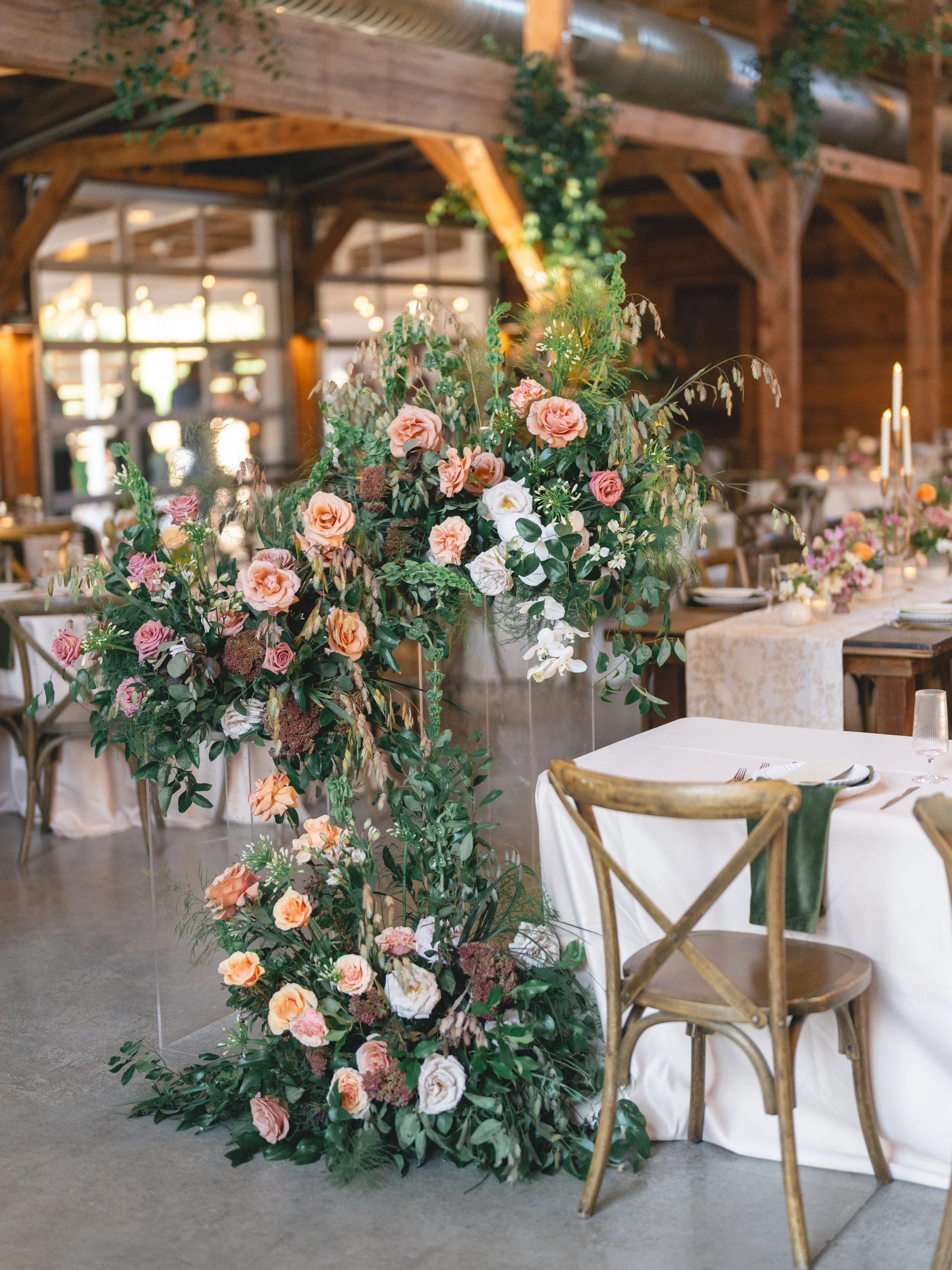  Floral installation of peach roses, white orchids and burgundy sedum spilling from a clear plinth beside the sweetheart table at The Addison Grove 