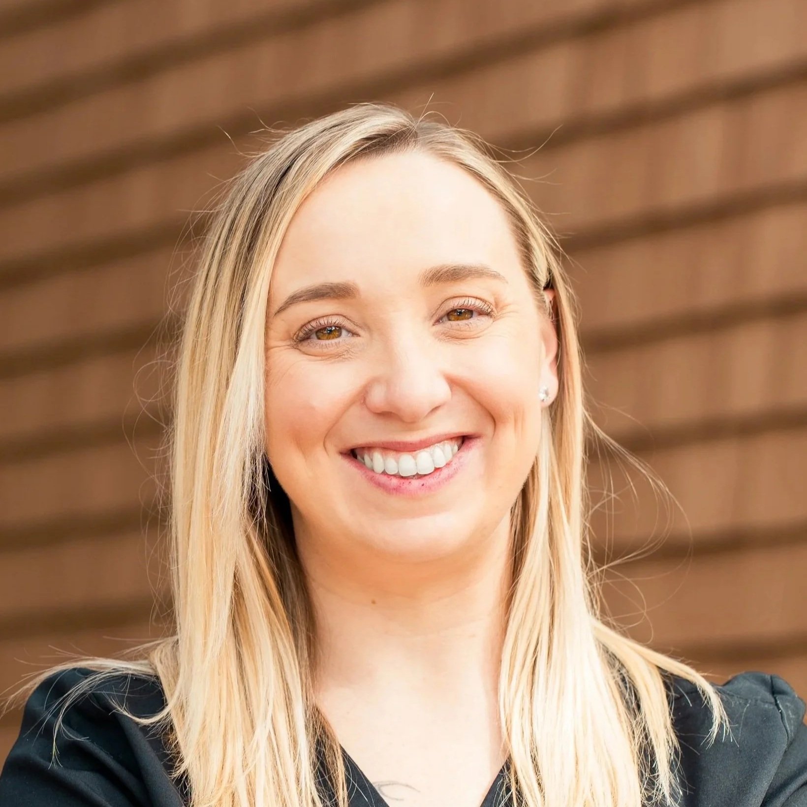 A smiling woman with long blonde hair and light skin, wearing a black top, standing in front of a wooden background.