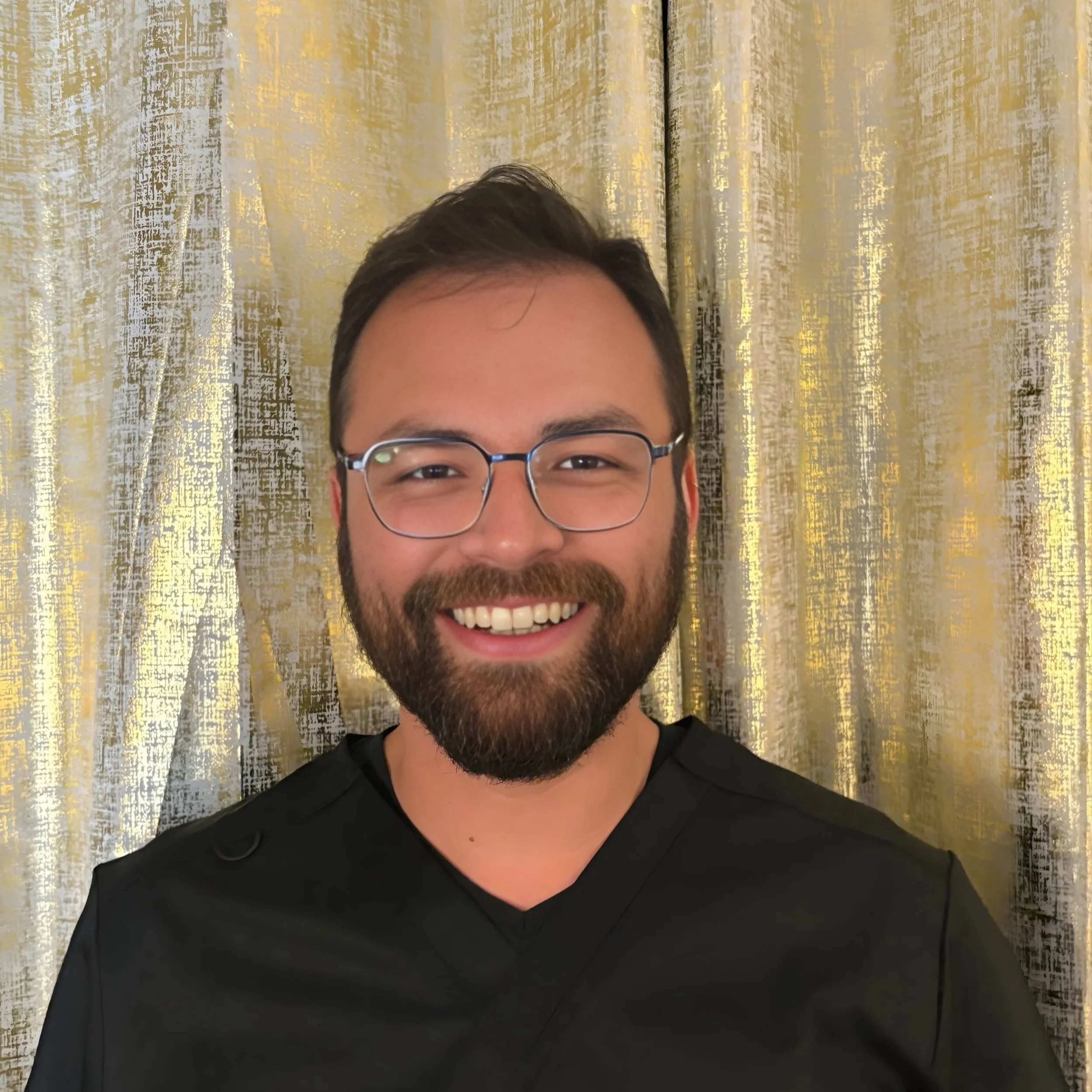 A smiling man with glasses, dark hair, and a beard, wearing a black shirt, standing in front of a gold and cream curtain.