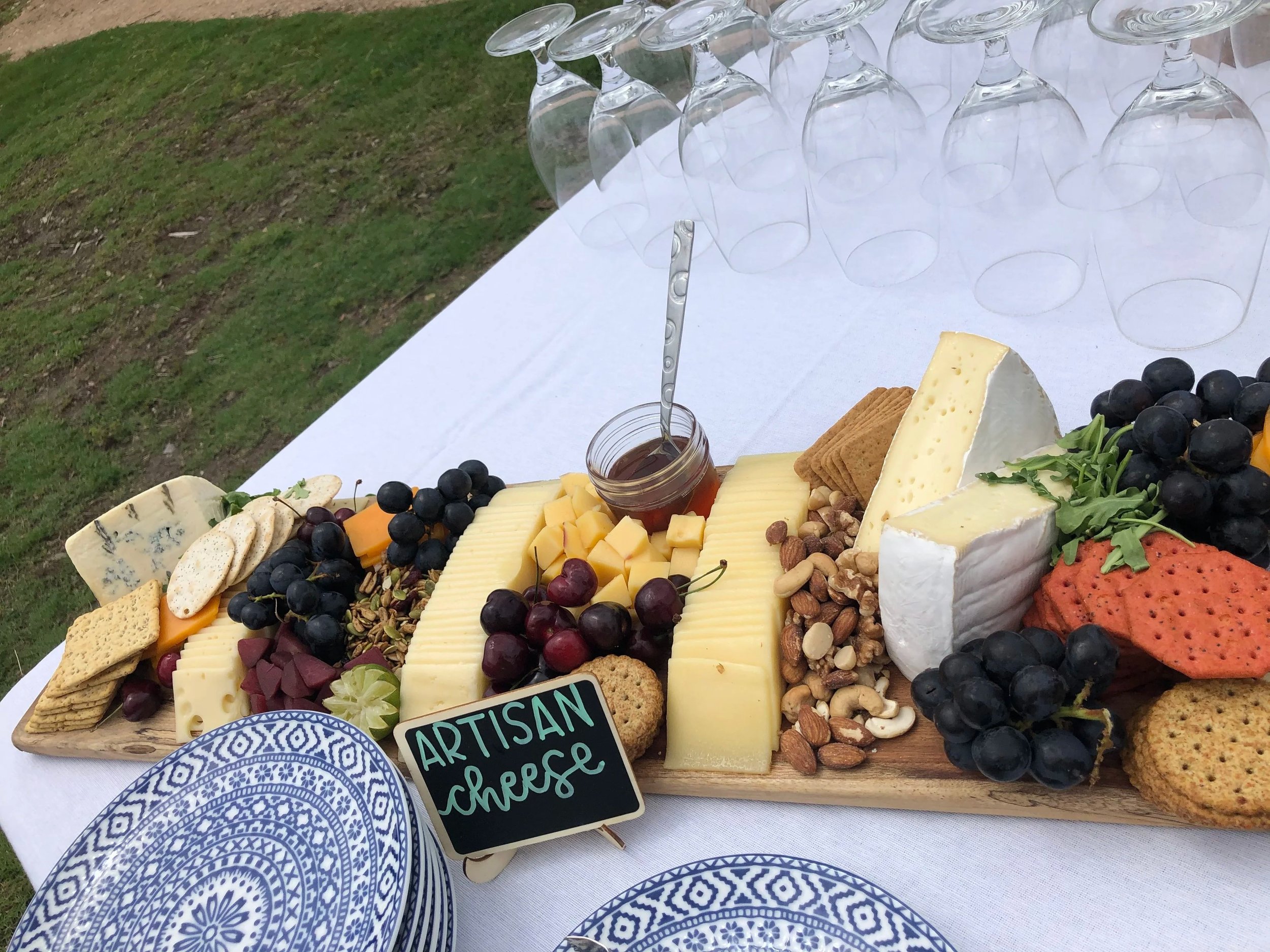 Cheese and charcuterie board with grapes, crackers, and honey, arranged on a wooden tray on a white tablecloth outdoors.
