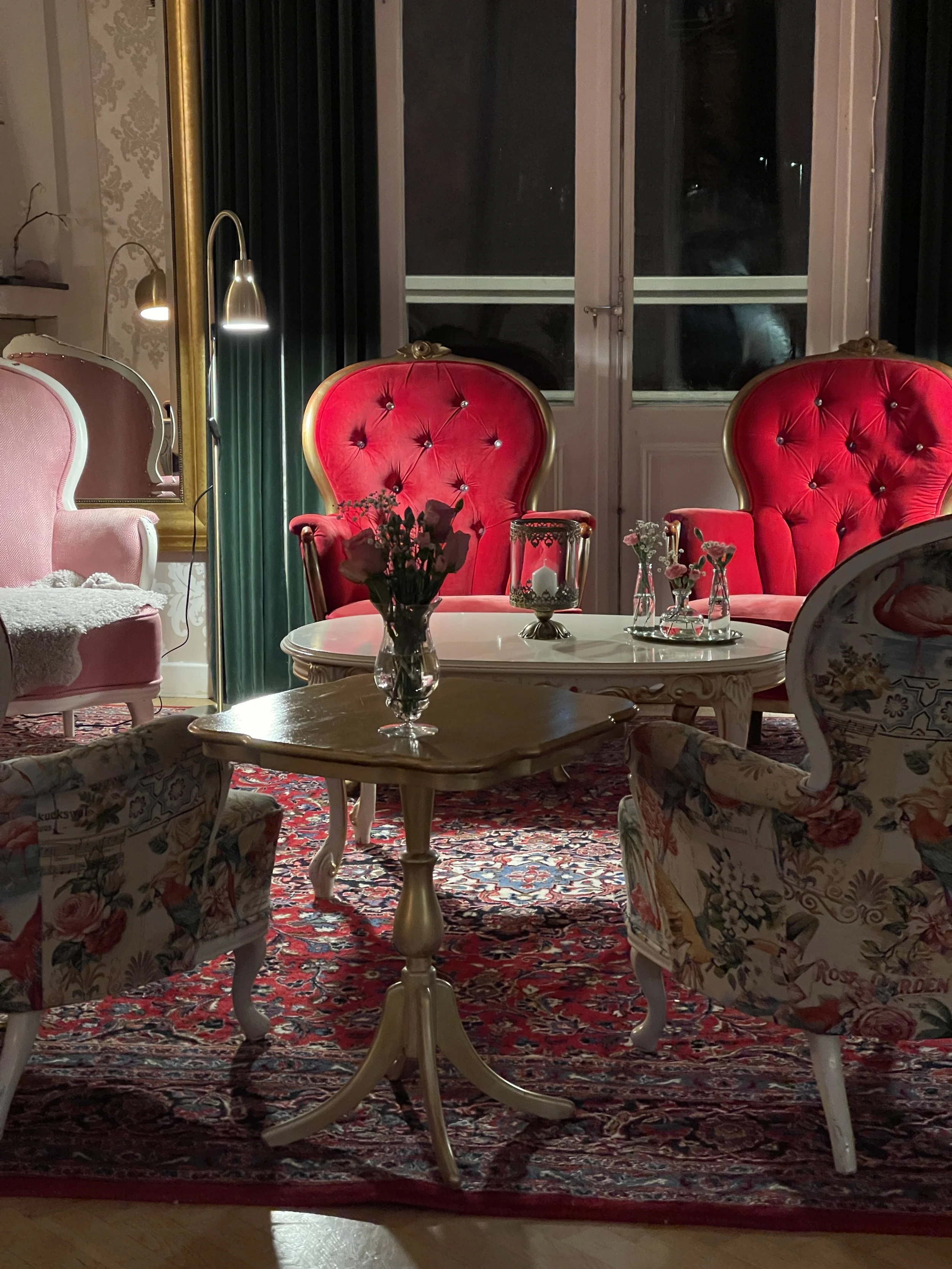 En stickhelg i mysigt vardagsrum med rosa och röda vintagefåtöljer, ett soffbord med blommor och ljuslyktor, en stor matta, en golvlampa och bakgrund med fönster och gardiner.