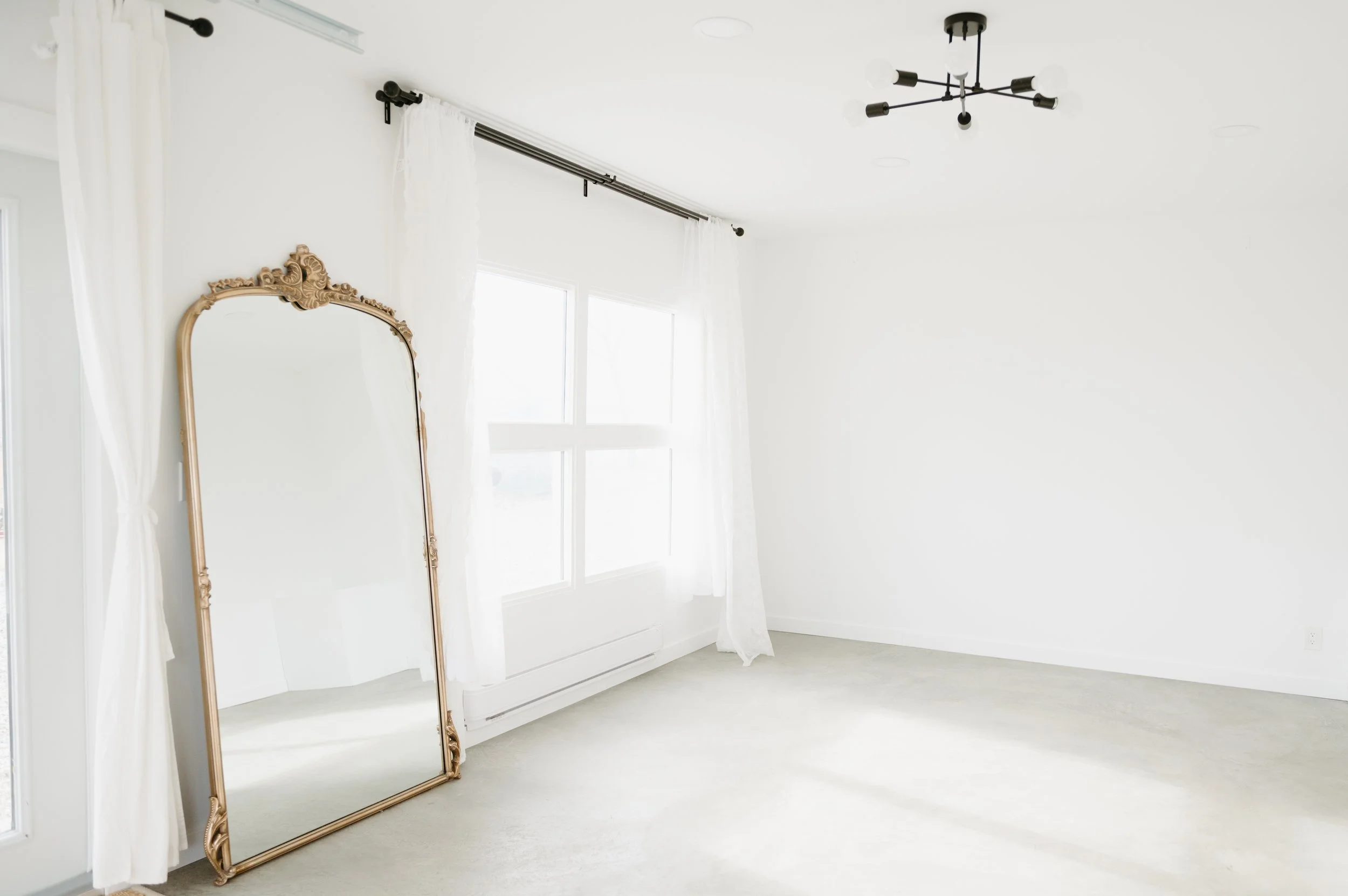 Empty room with white walls, large windows with white curtains, a gold ornate mirror leaning against the wall, and a modern black chandelier on the ceiling.