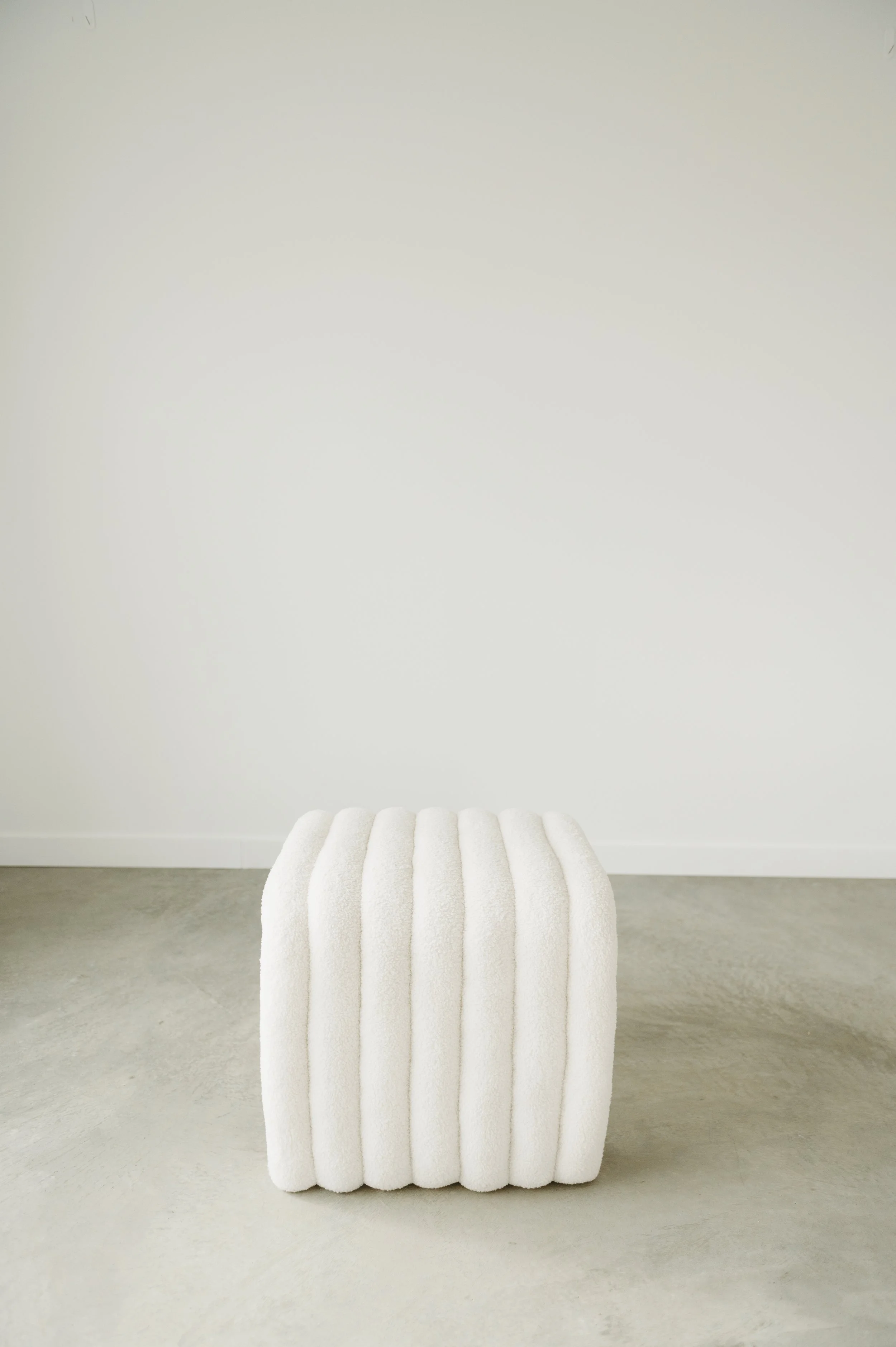 A white, textured, plush cubic stool or ottoman placed on a light gray concrete floor against a plain white wall.