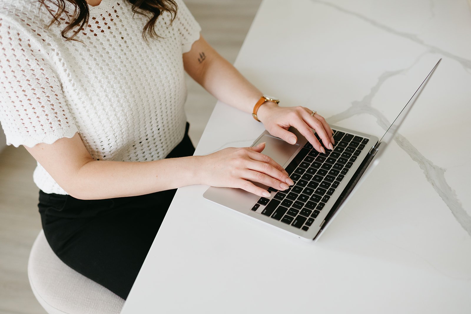 A woman wearing a white knitted top and black pants sitting at a white desk, using a silver laptop.