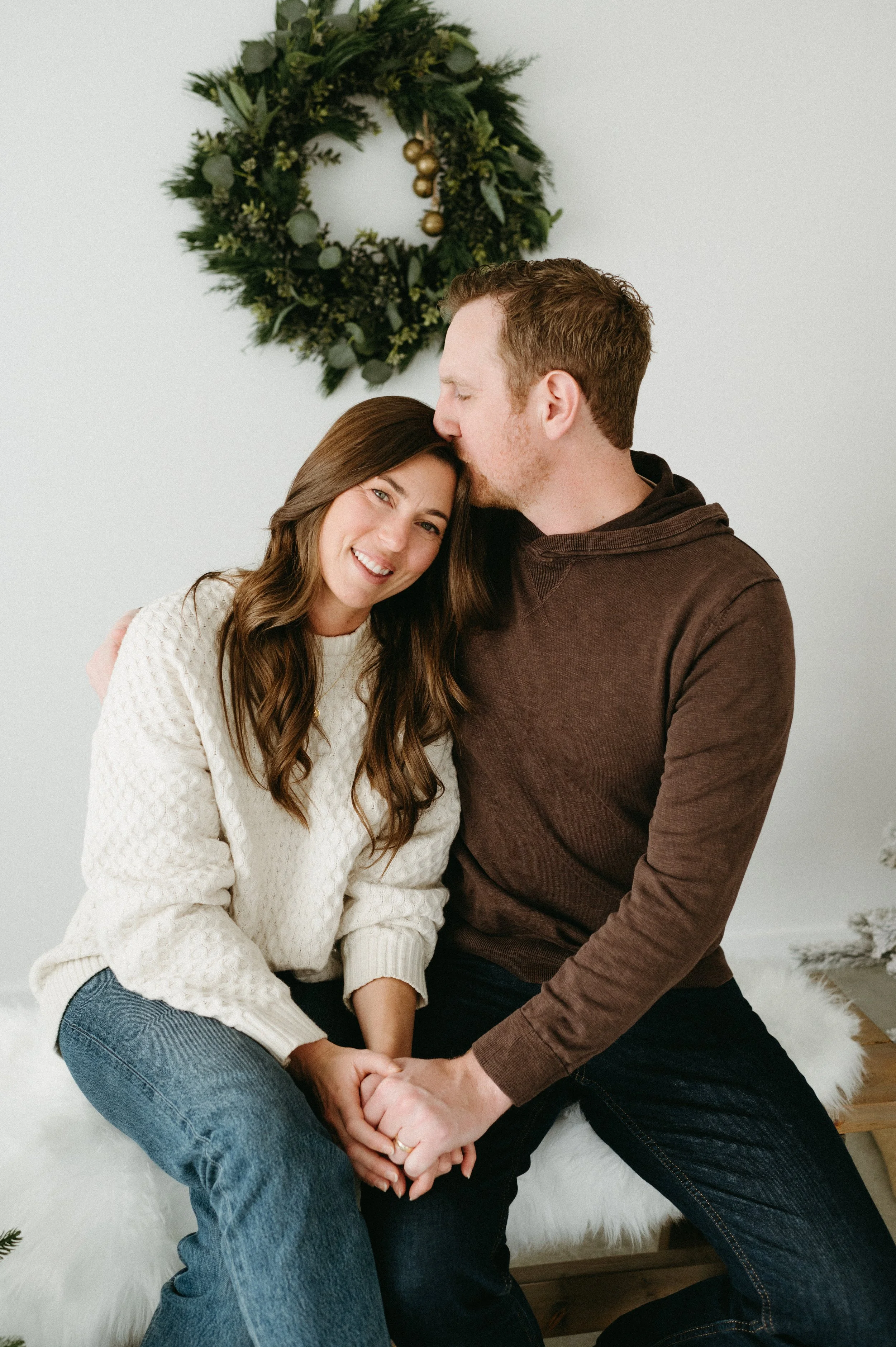 A smiling woman and a man sitting close together, holding hands, with a Christmas wreath on the wall behind them.