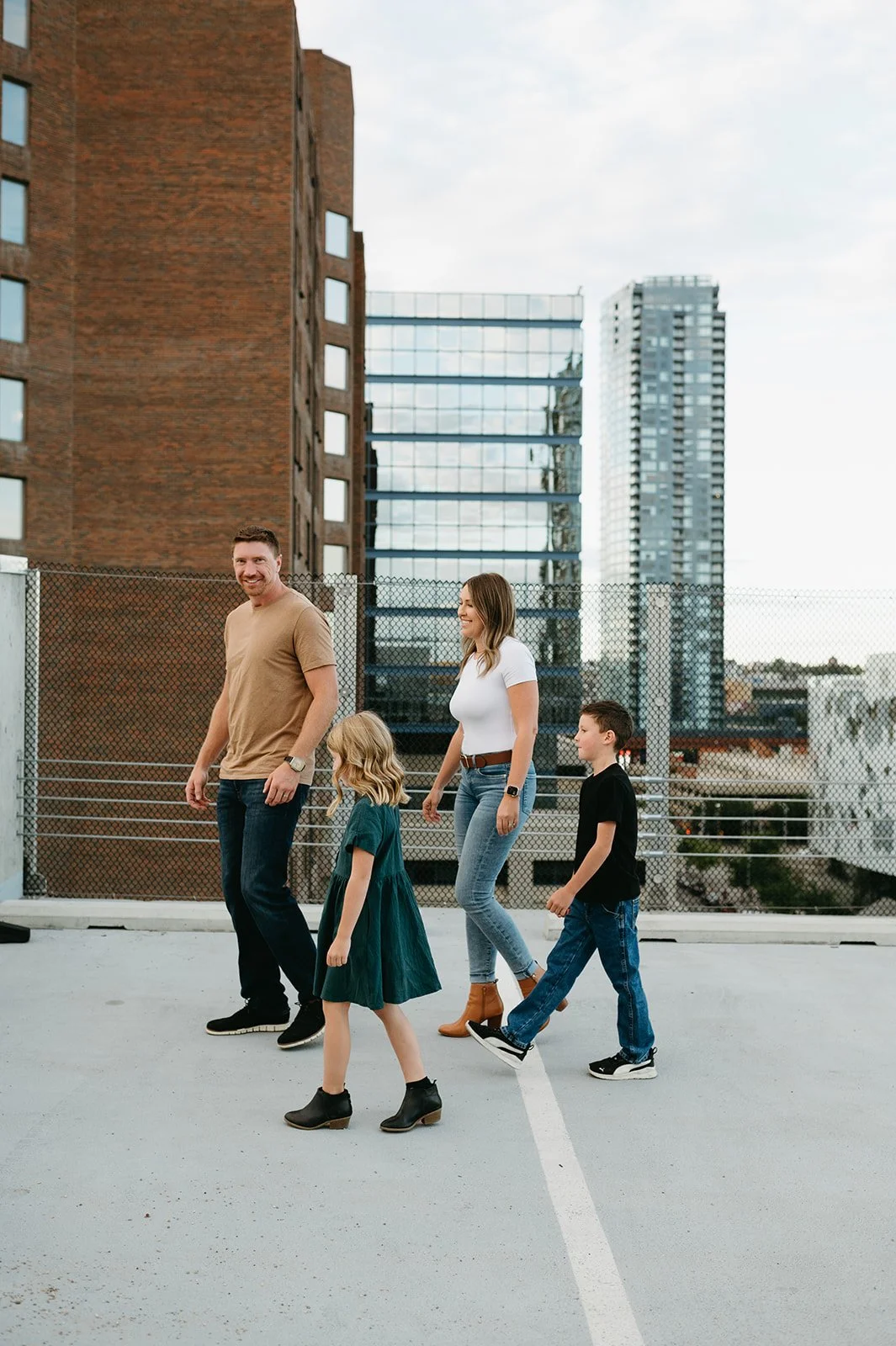 A family walking in an outdoor parking lot with a cityscape background, including buildings and a fence.