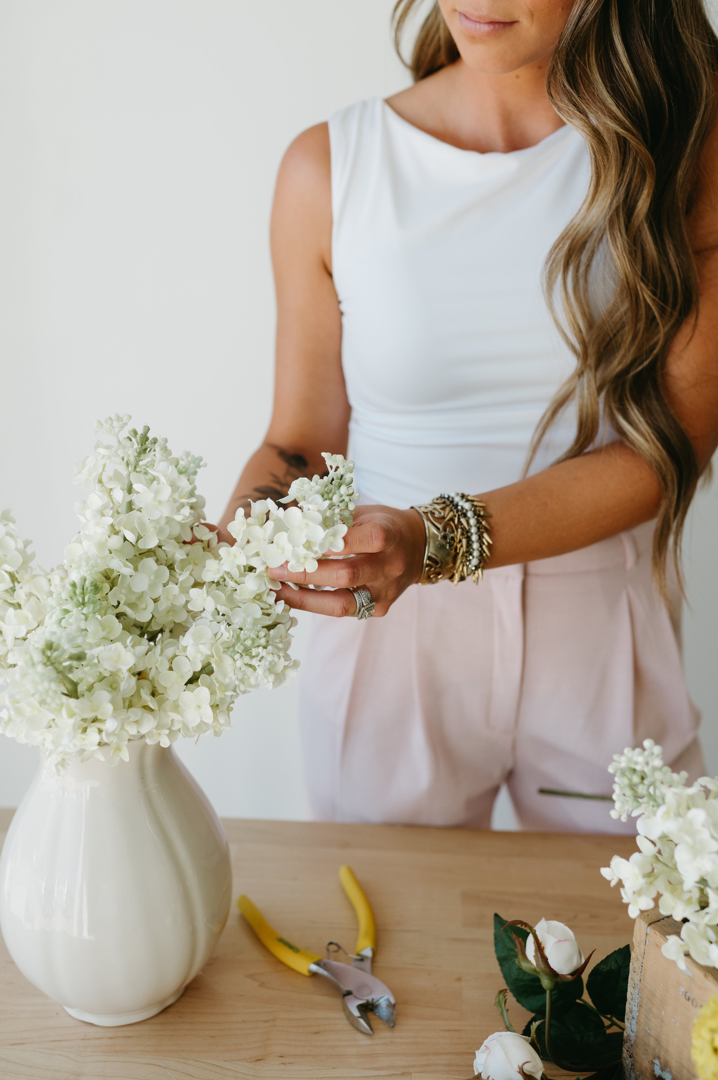 Woman arranging white flowers in a ceramic vase, wearing a white sleeveless top and light pink pants, with jewelry on her wrist, on a wooden table with yellow pruning shears and additional flowers.