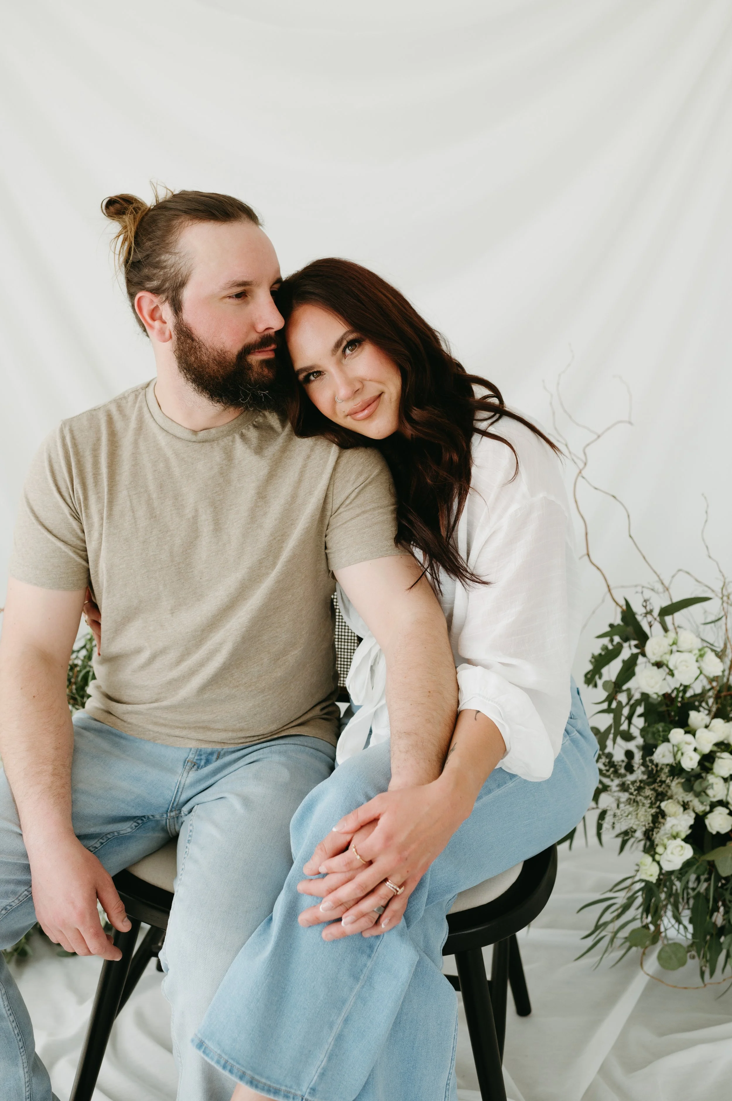 A couple sitting closely together on a black chair, surrounded by white flowers and greenery, with a white backdrop.