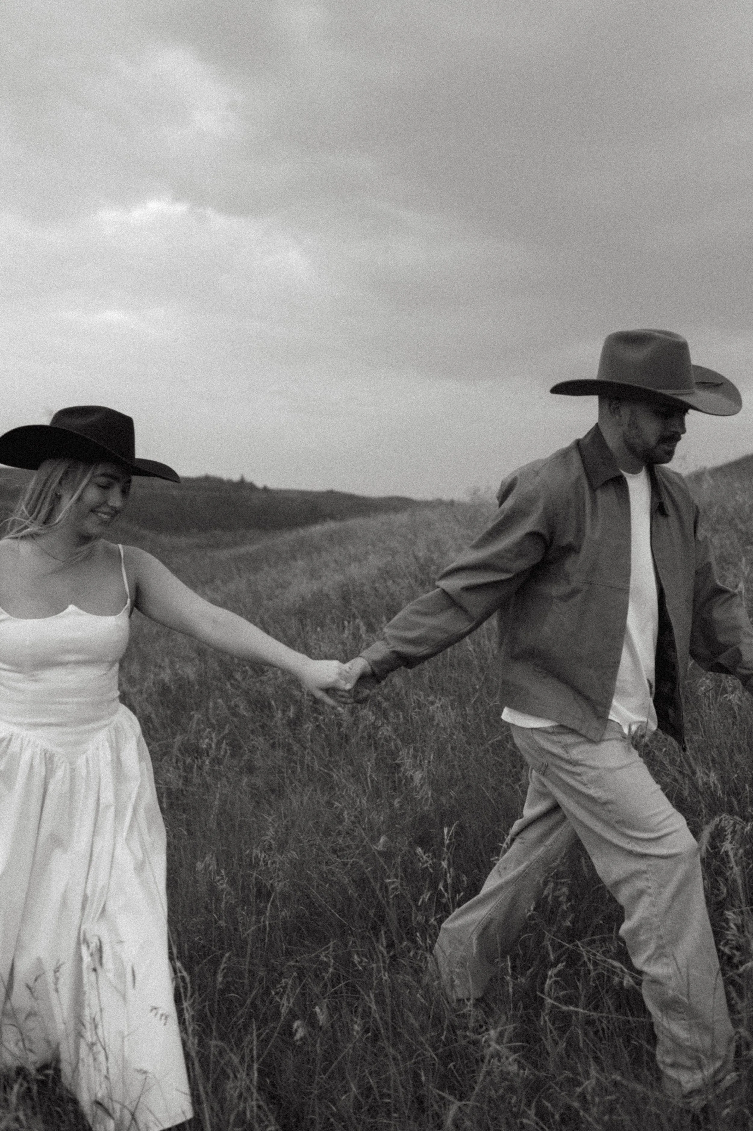 A black and white photo of a couple holding hands while walking through a grassy field, both wearing cowboy hats. The woman is in a white dress, and the man is in a jacket and pants.