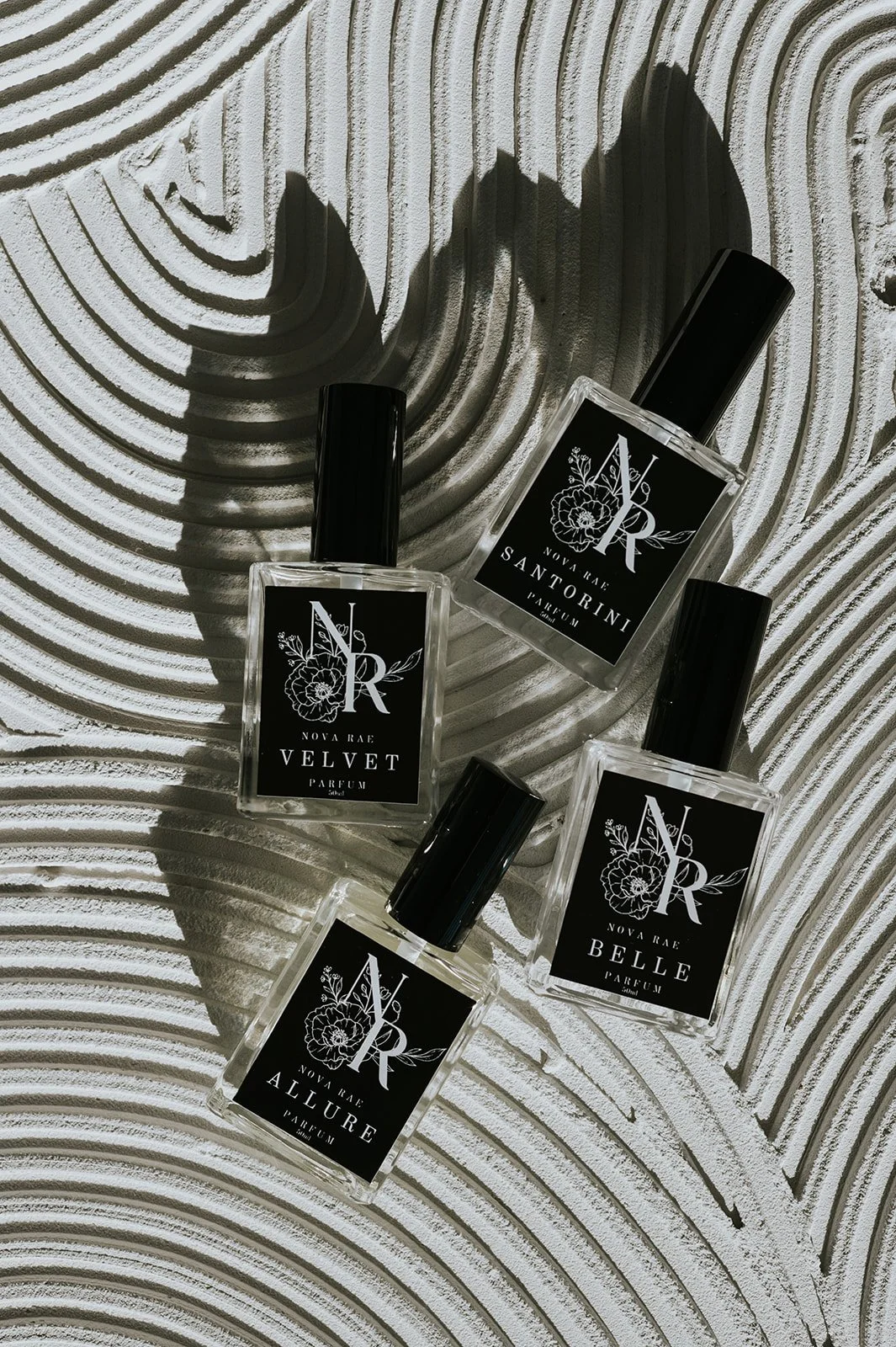 Four bottles of Nova Rae perfume, labeled Velvet, Santorini, Belle, and Allure, on a textured, white, wavy background.