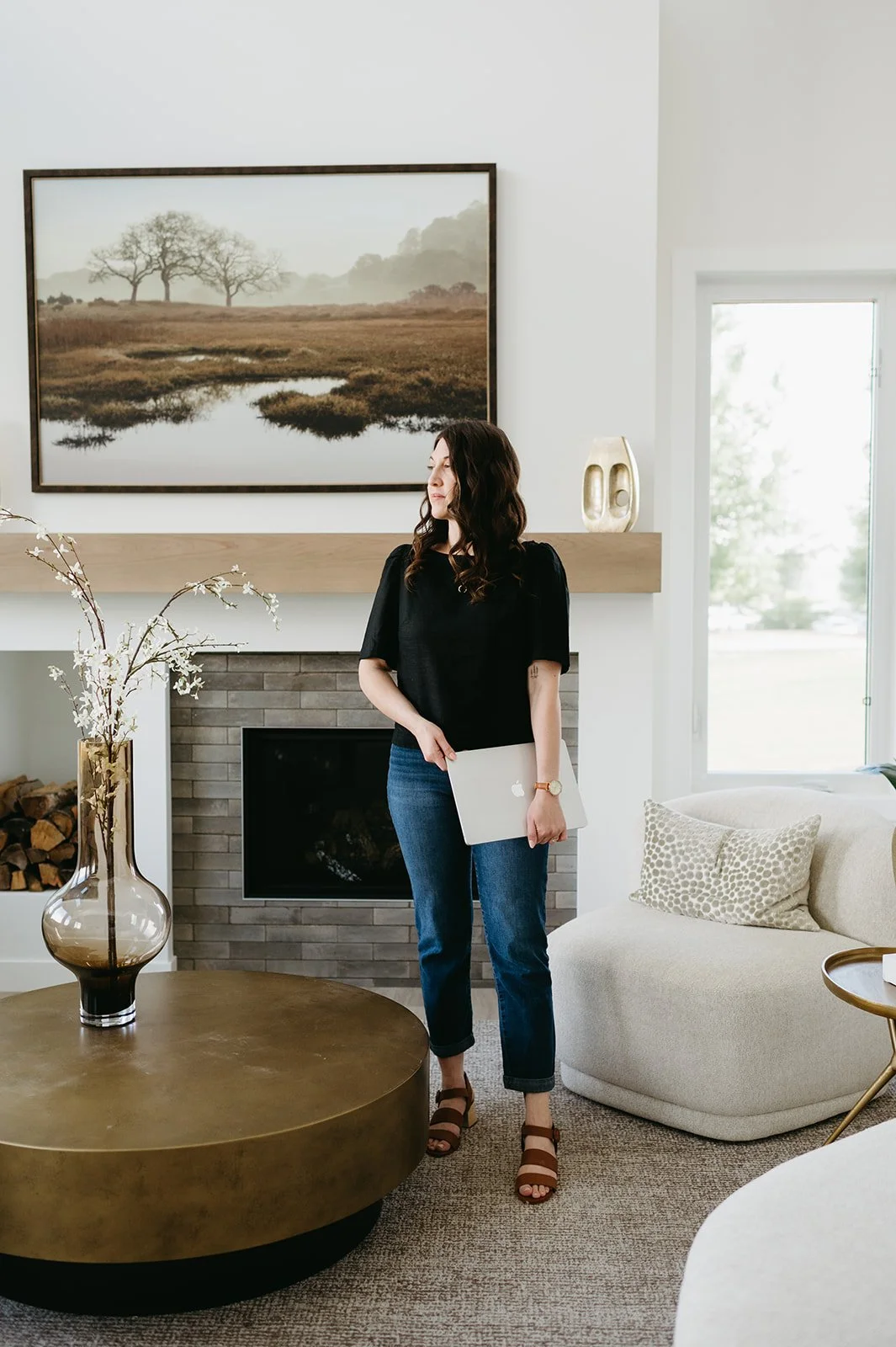A woman with brown hair, wearing a black top, blue jeans, and brown sandals, holding a silver laptop, standing in a modern living room near a fireplace with a large landscape painting above it.