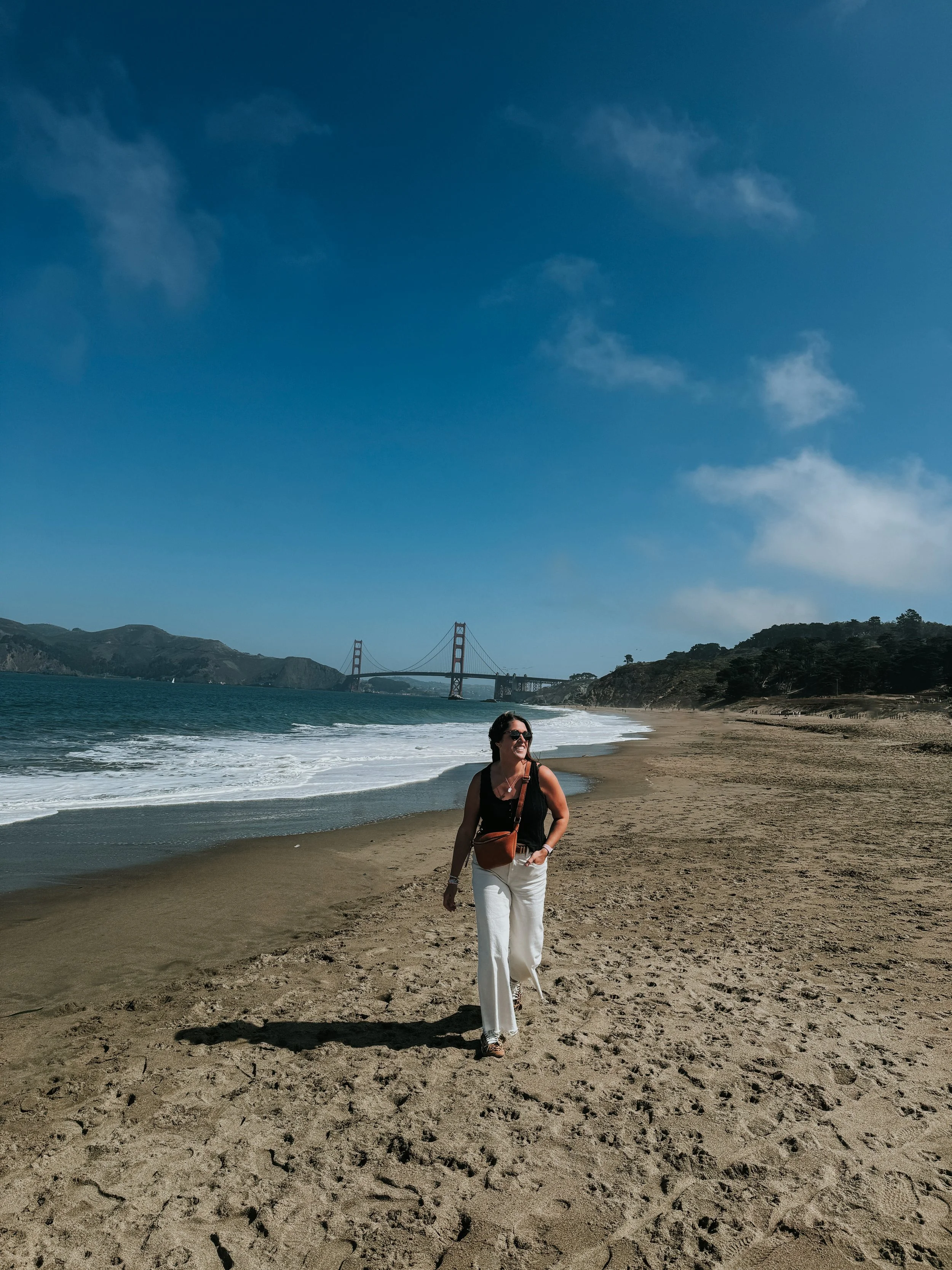 A woman walking on a beach near the ocean with the Golden Gate Bridge in the background under a clear blue sky.