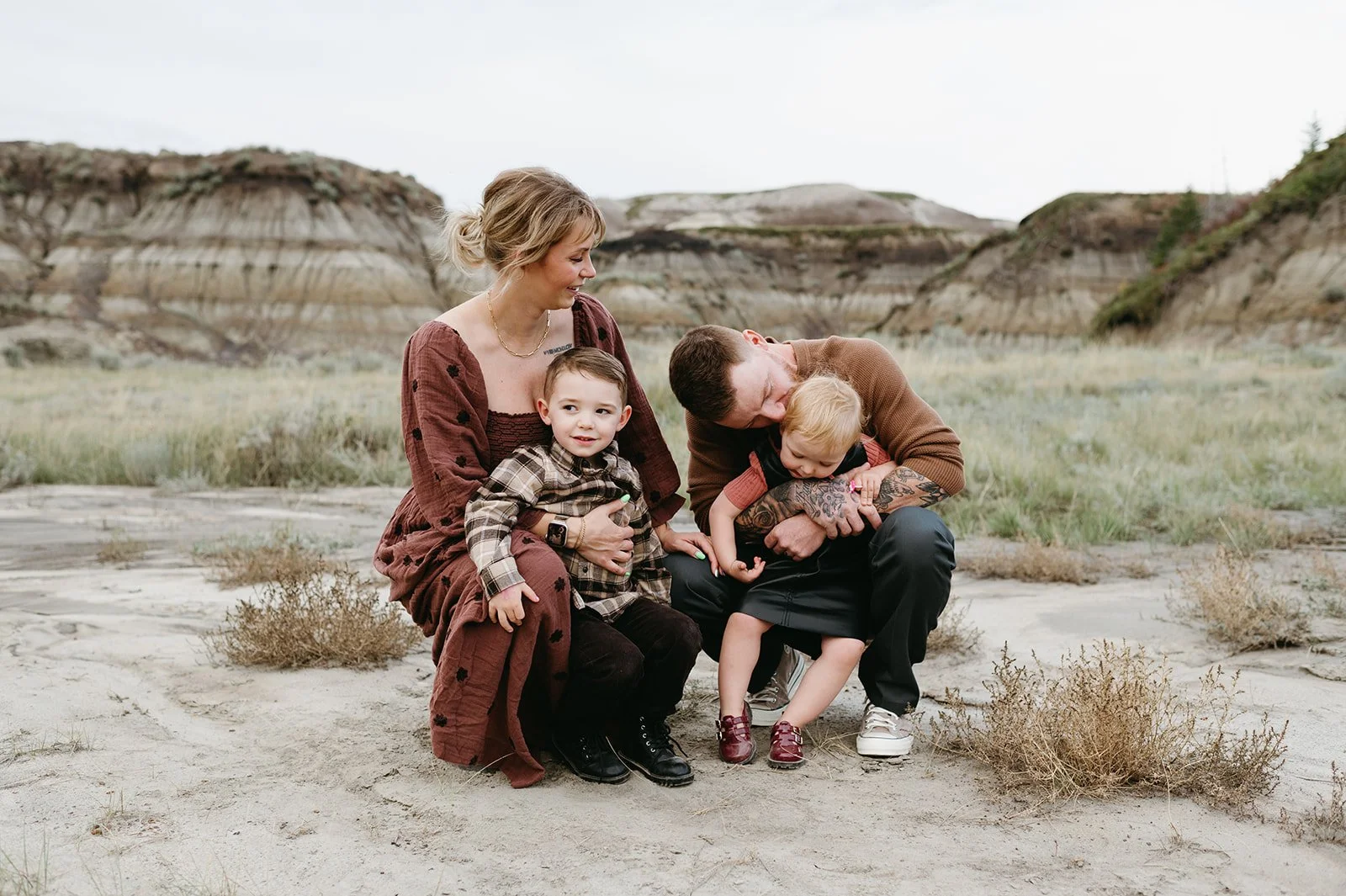 A family of four sitting outdoors on the ground in a desert landscape with layered rock formations in the background. The mother has blonde hair and is wearing a brown dress, holding a young boy in a plaid shirt on her lap. The father, with tattoos, 