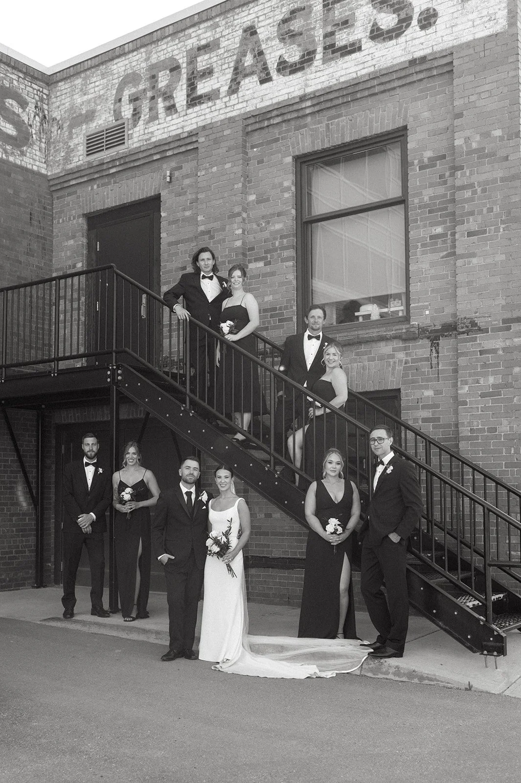 Black and white photo of a wedding party with eight people, some on stairs and some on the ground, in front of a brick building with a large window and a faded sign that says 'GREAT EASES'.