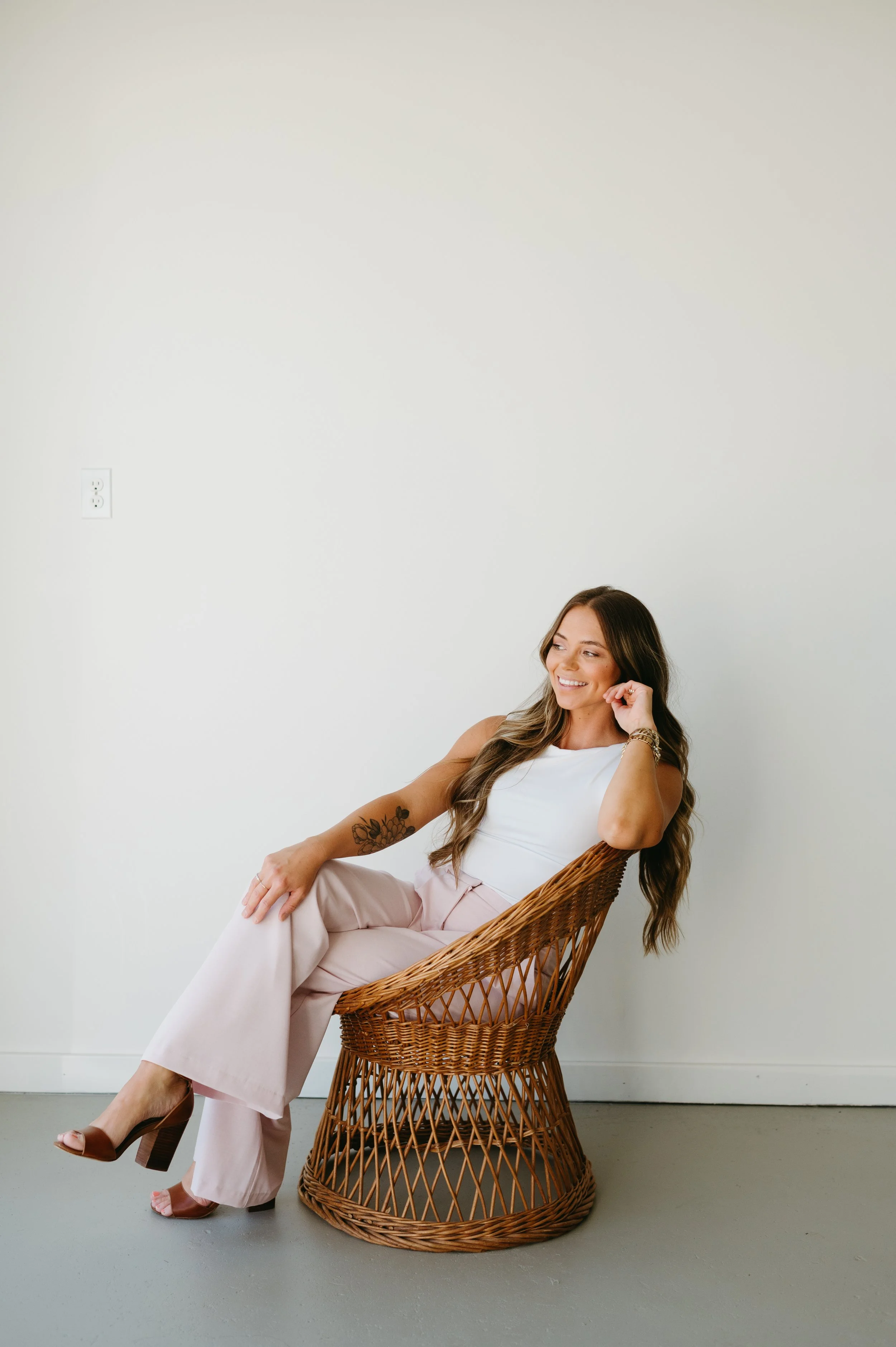 A woman with long brown hair, wearing a white sleeveless top, pink wide-leg pants, and brown heels, sitting in a wicker chair against a plain white wall, smiling and looking to the side.