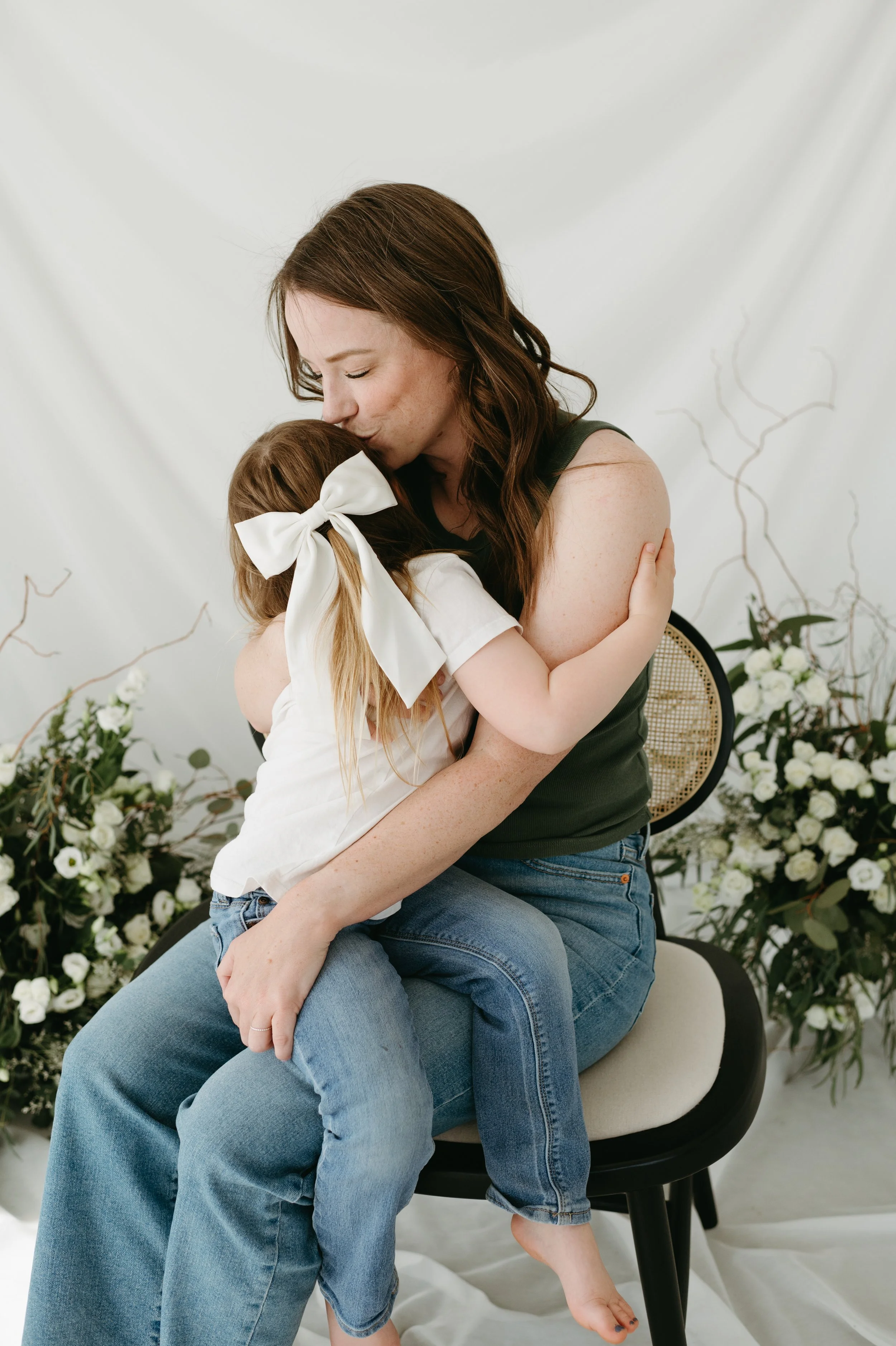 A woman with long brown hair hugging a girl with long blonde hair tied with a large white bow, surrounded by floral arrangements.