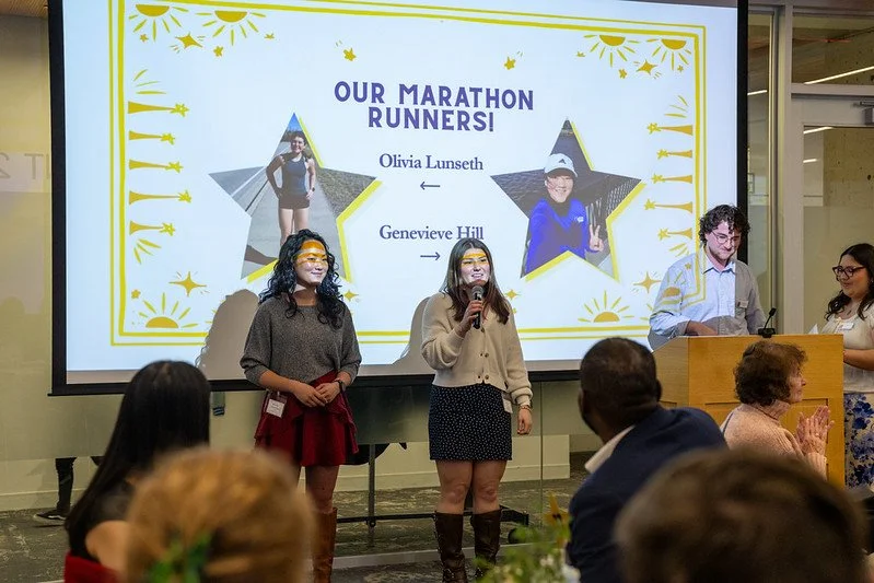A group of people standing in front of a screen displaying a "Thank You to our Boston Marathon Runners" message at an indoor event. One person holds a microphone, and a projector shows images and text. The room appears to be a conference or event space with flowers and artwork visible.