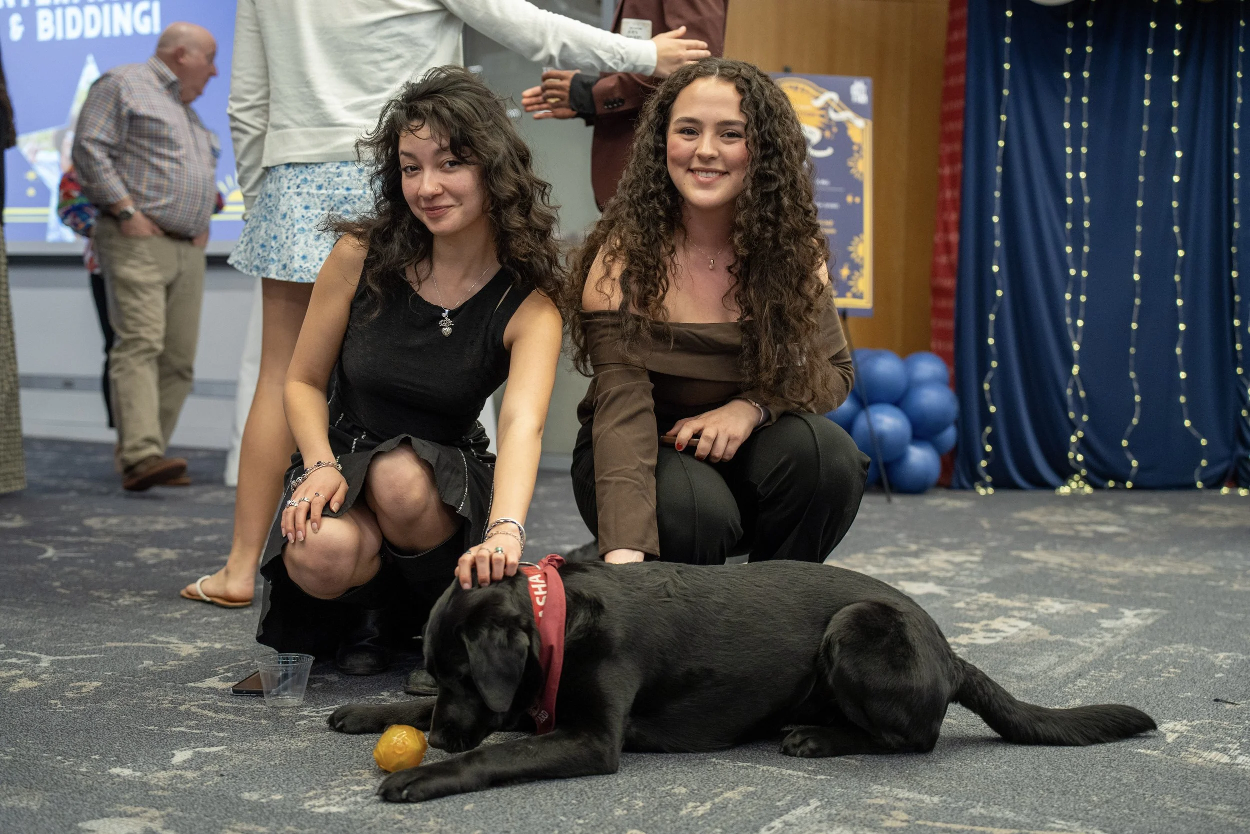 Two young women kneel next to a black Labrador puppy with a red collar, playing with a yellow toy ball on the floor at an indoor event. The background includes a blue curtain, a group of balloons, and a screen with writing.