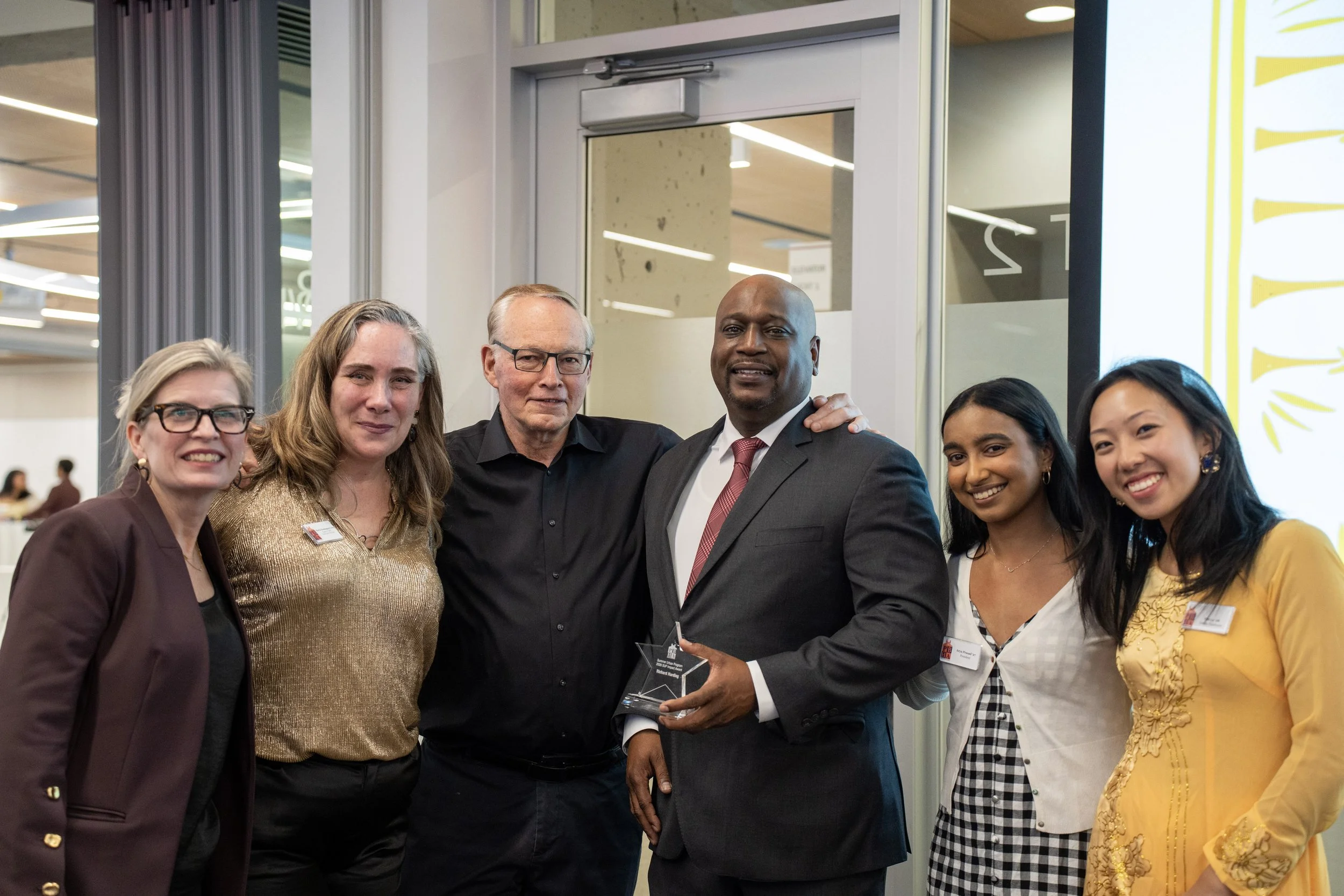 Group photo at an award event, featuring a presentation screen with 'Awarded to Ekua Holmes,' showing people standing together smiling, with flowers in foreground.