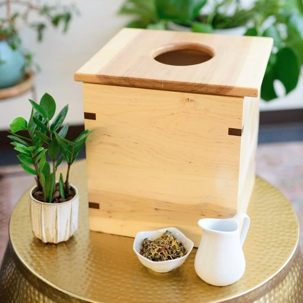 A wooden box with a round opening on top sits on a round, gold-colored table alongside a potted plant, a small white bowl with herbs, and a tiny white pitcher.