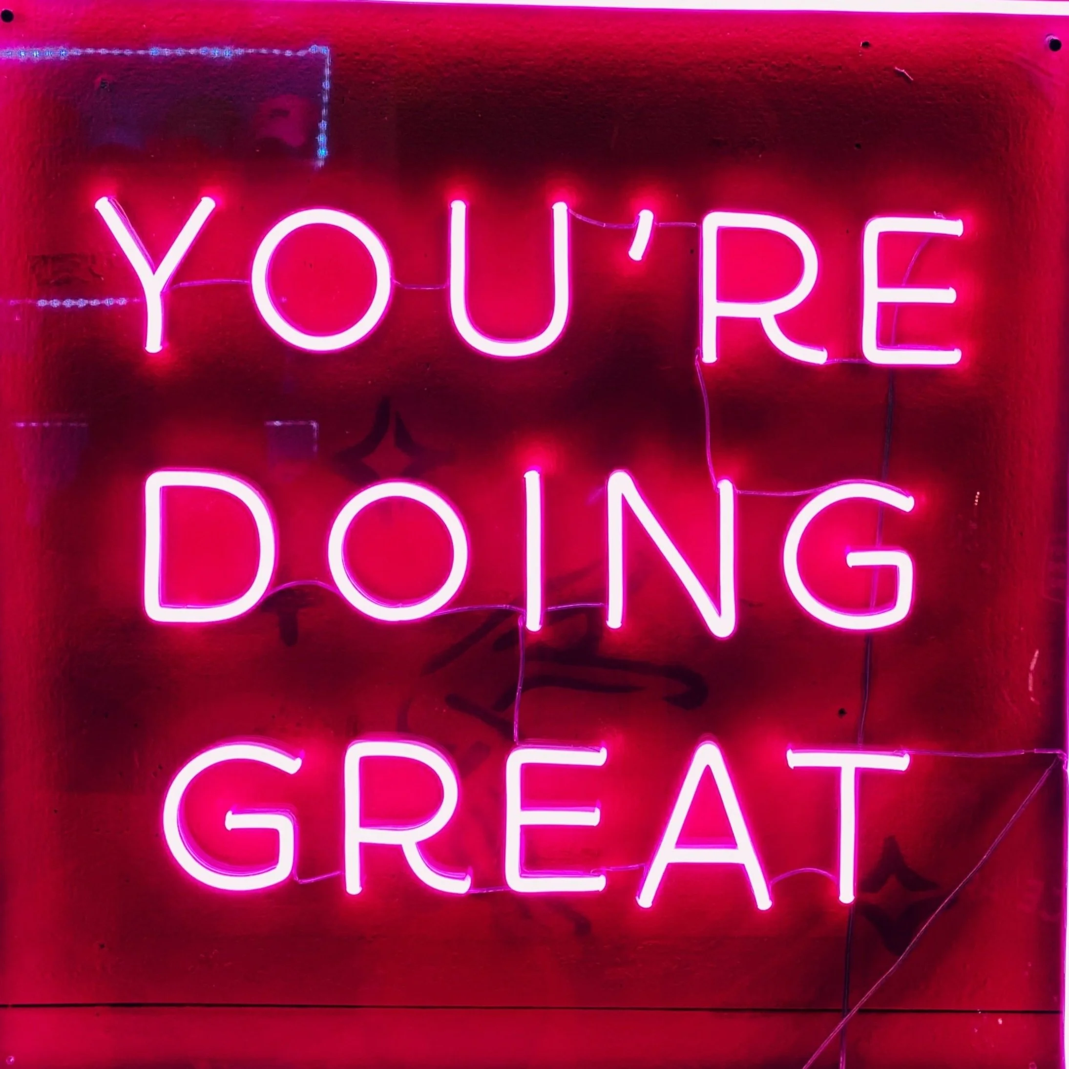 Pink neon sign displaying the phrase 'You're doing great' against a red background.