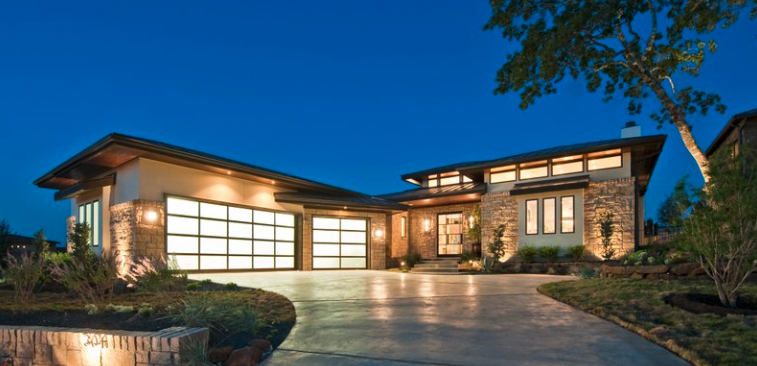 Modern residential house with large windows, stone facade, and a landscaped front yard at dusk.