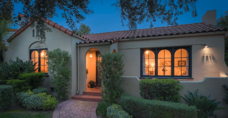 Front view of a cozy house with a red tiled roof, surrounded by green bushes and trees, during dusk with lights on inside.