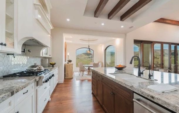 Open concept kitchen with white cabinets, granite countertops, and a central island, adjacent to a dining area with a chandelier, large windows, and views of green hills.