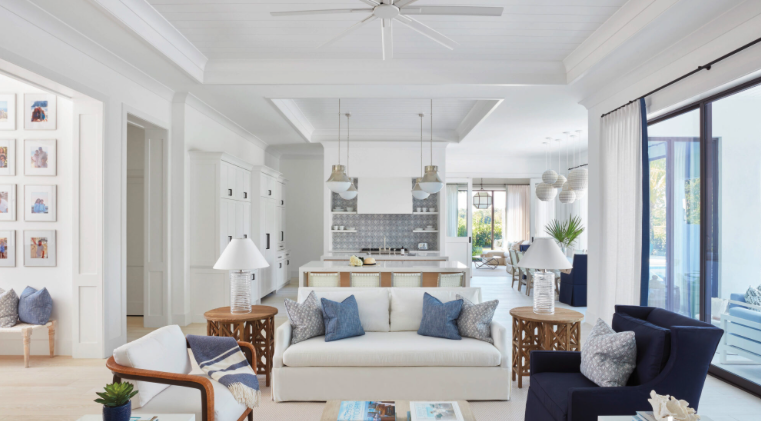 Bright open-concept living room with white sofa, navy armchair, and side tables, leading to a modern kitchen and dining area with large windows and sliding glass door.