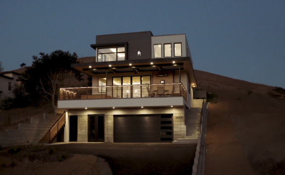Modern multi-story house on a hillside at dusk with exterior lighting, large windows, and a garage.