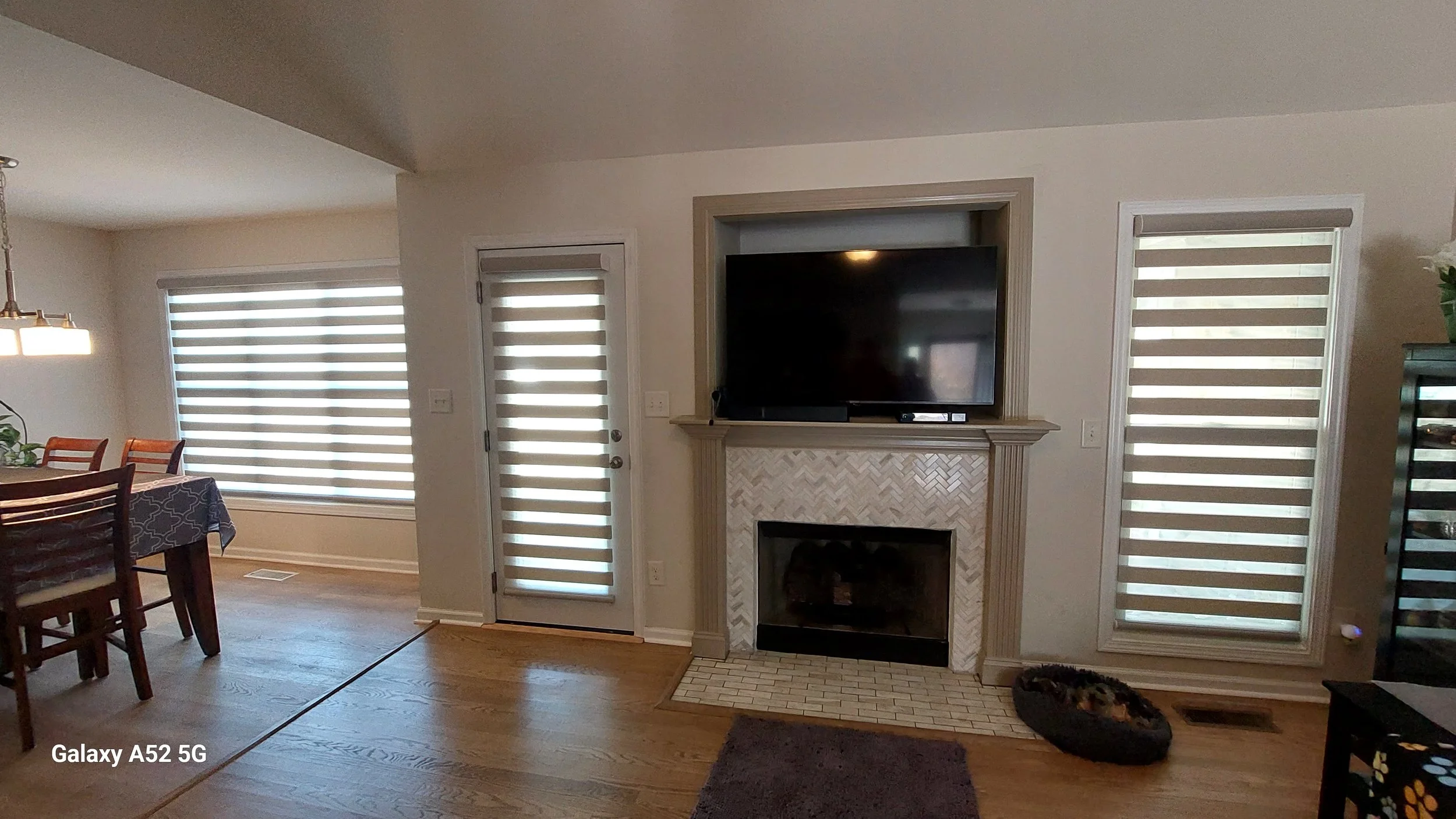Neutral banded shades installed on large windows and a glass patio door in a bright open-concept living area with wooden floors, a fireplace, and an adjacent dining room.