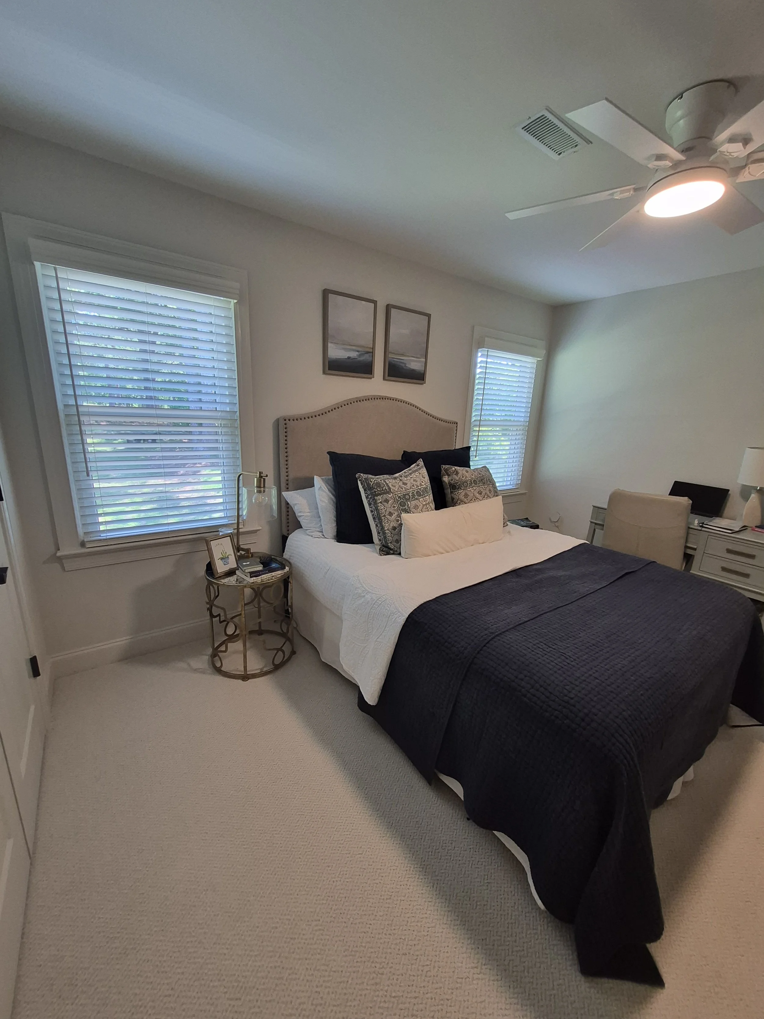 White faux wood blinds installed on windows in a modern bedroom with light gray walls, carpeted floors, and a dark blue duvet.