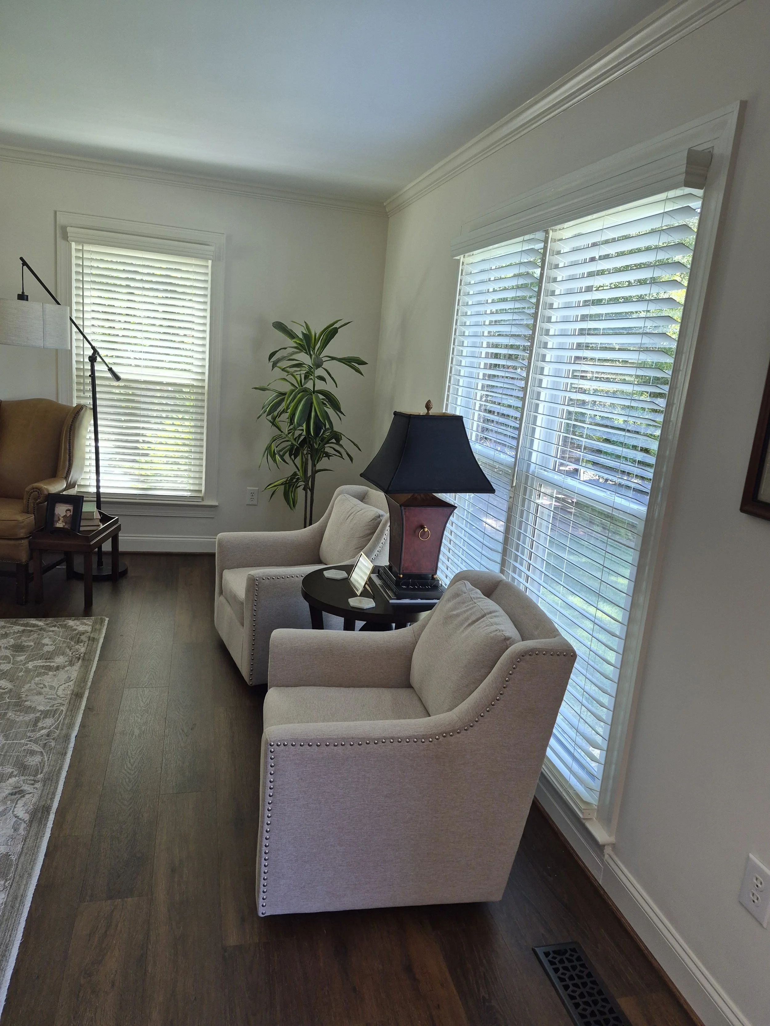 White wood blinds installed on multiple windows in a sunlit living room with dark hardwood floors and beige armchairs.
