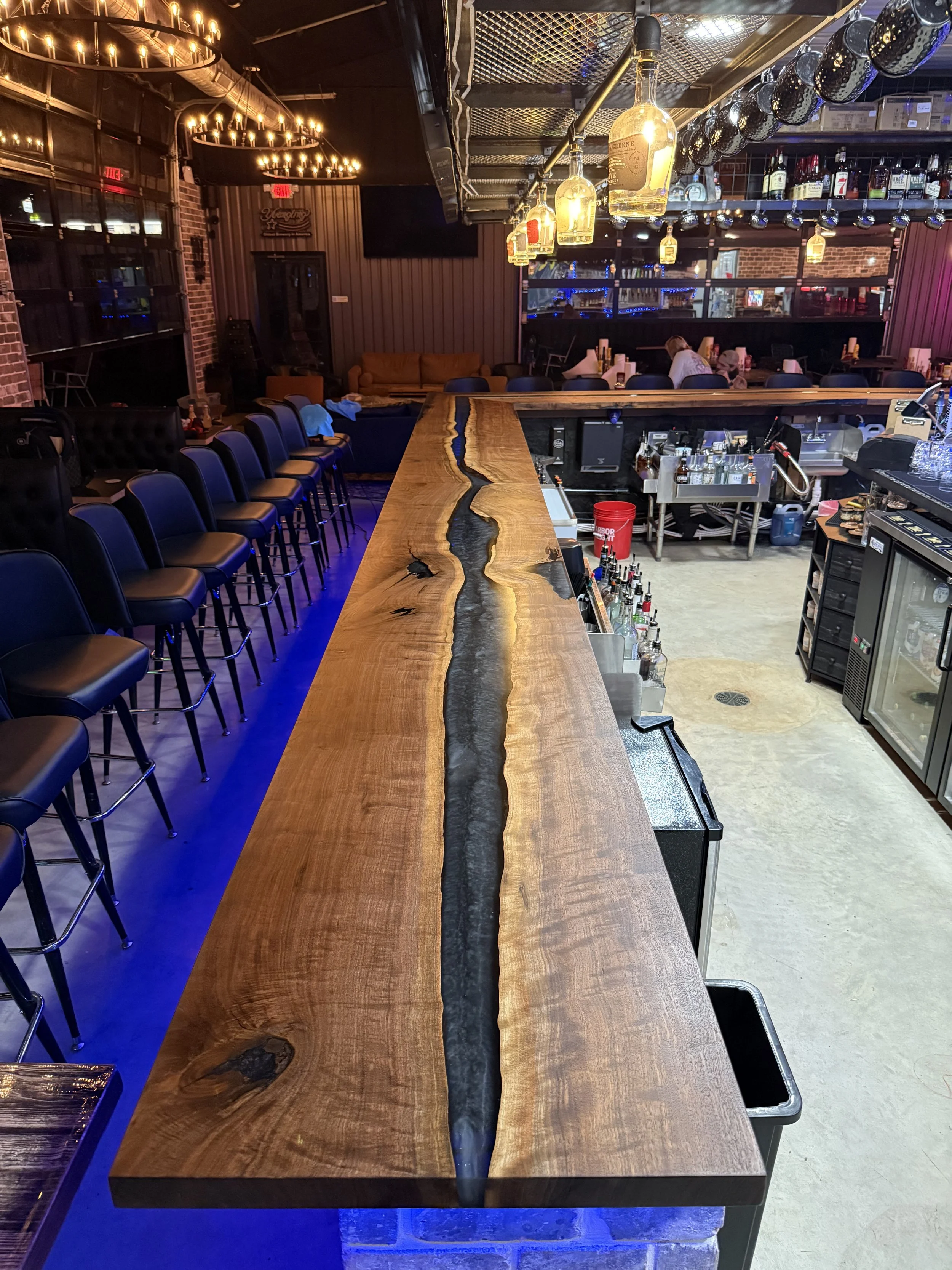 The image shows the interior of a bar or restaurant with a long wooden bar counter, bar stools, hanging wine bottle lights, and seating areas in the background.