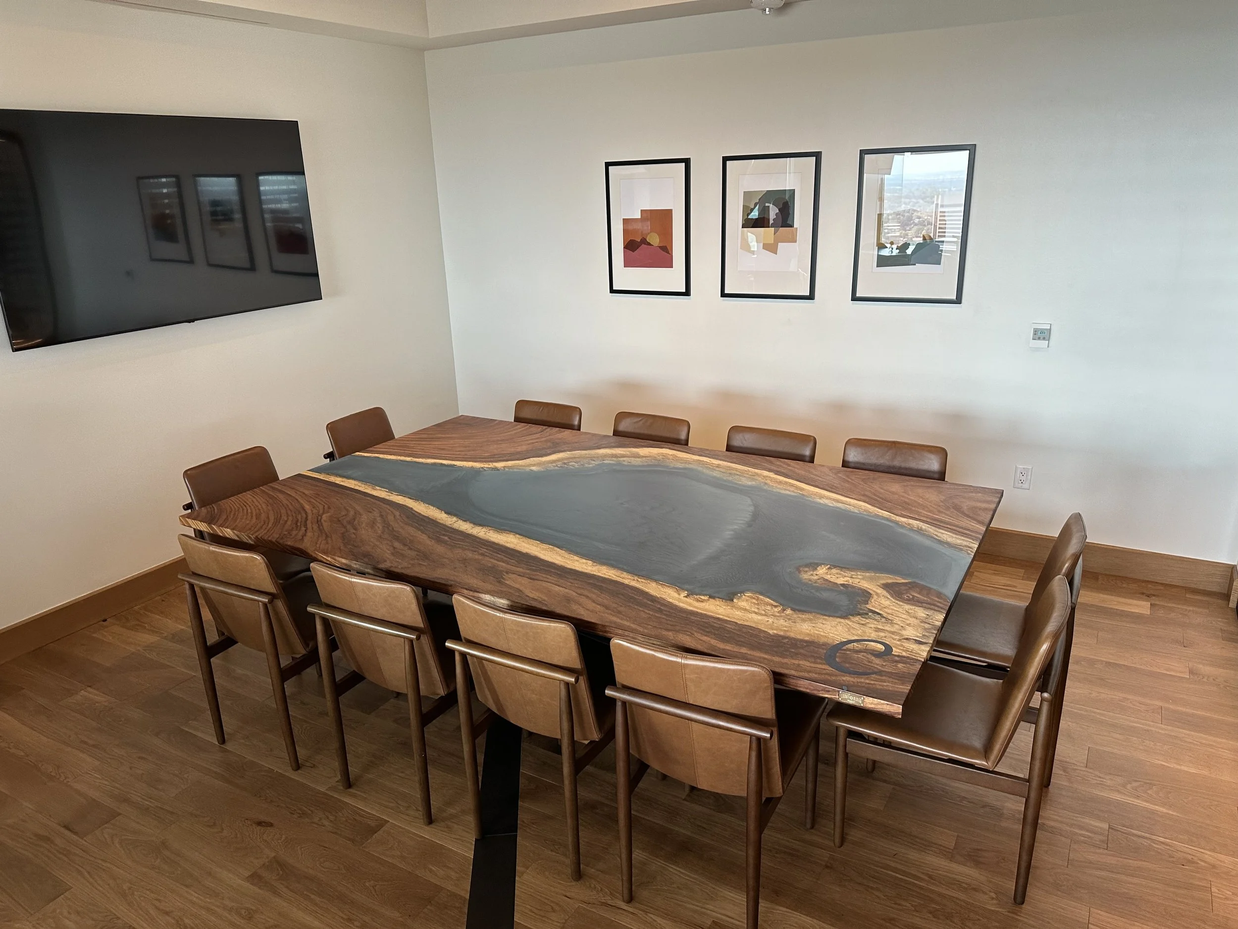 A modern conference room with a large, rectangular wooden table featuring a black and gold resin inlay. There are nine brown leather chairs around the table, and a flat-screen TV mounted on the wall. Three framed abstract art pieces hang on the wall, and the room has wooden flooring.