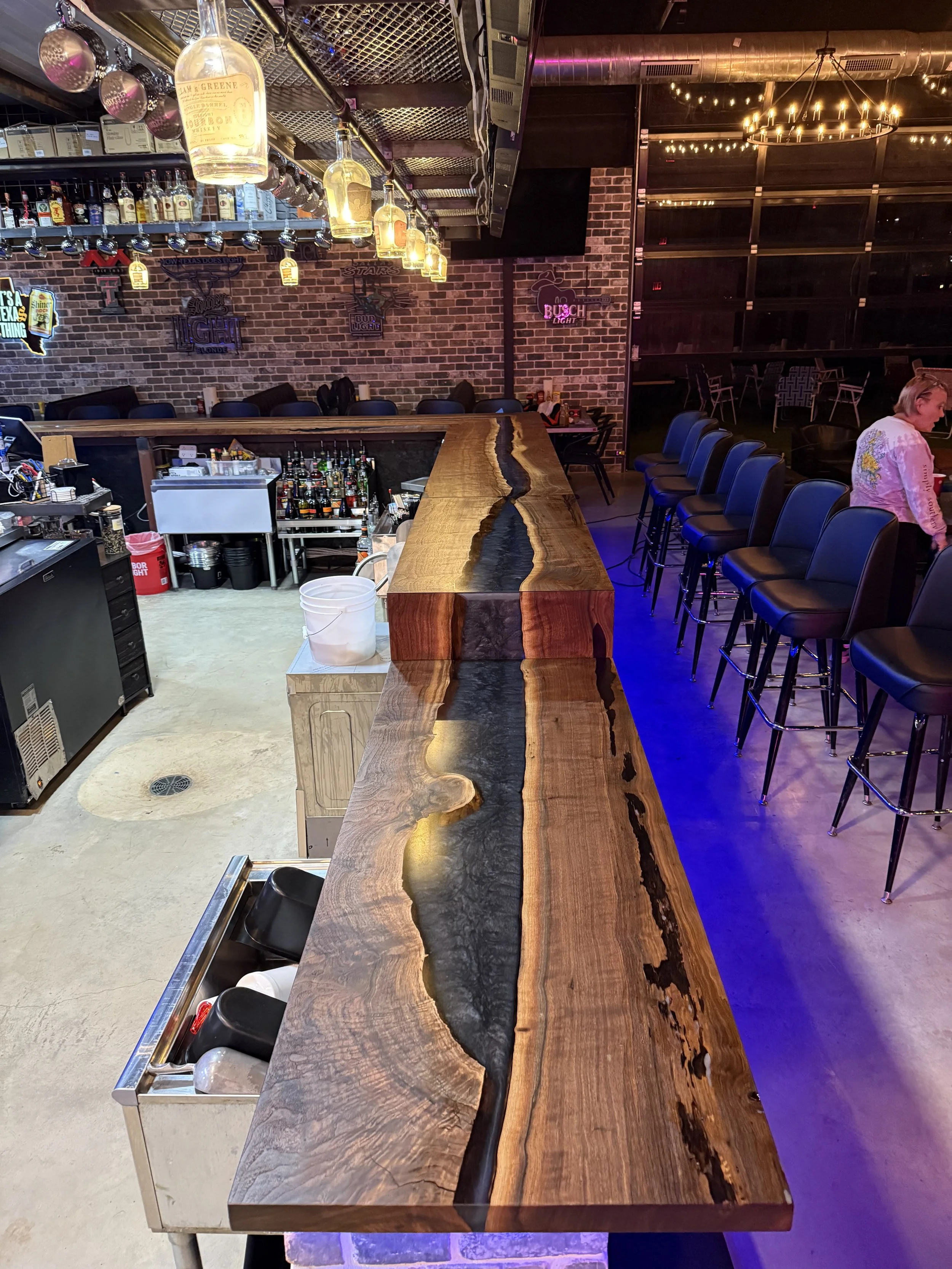 Empty bar counter made from a thick wood slab with a black resin river running through it, set in a bar or lounge with chairs and bottles of alcohol behind it, decorated with hanging lights and signs on the brick wall.