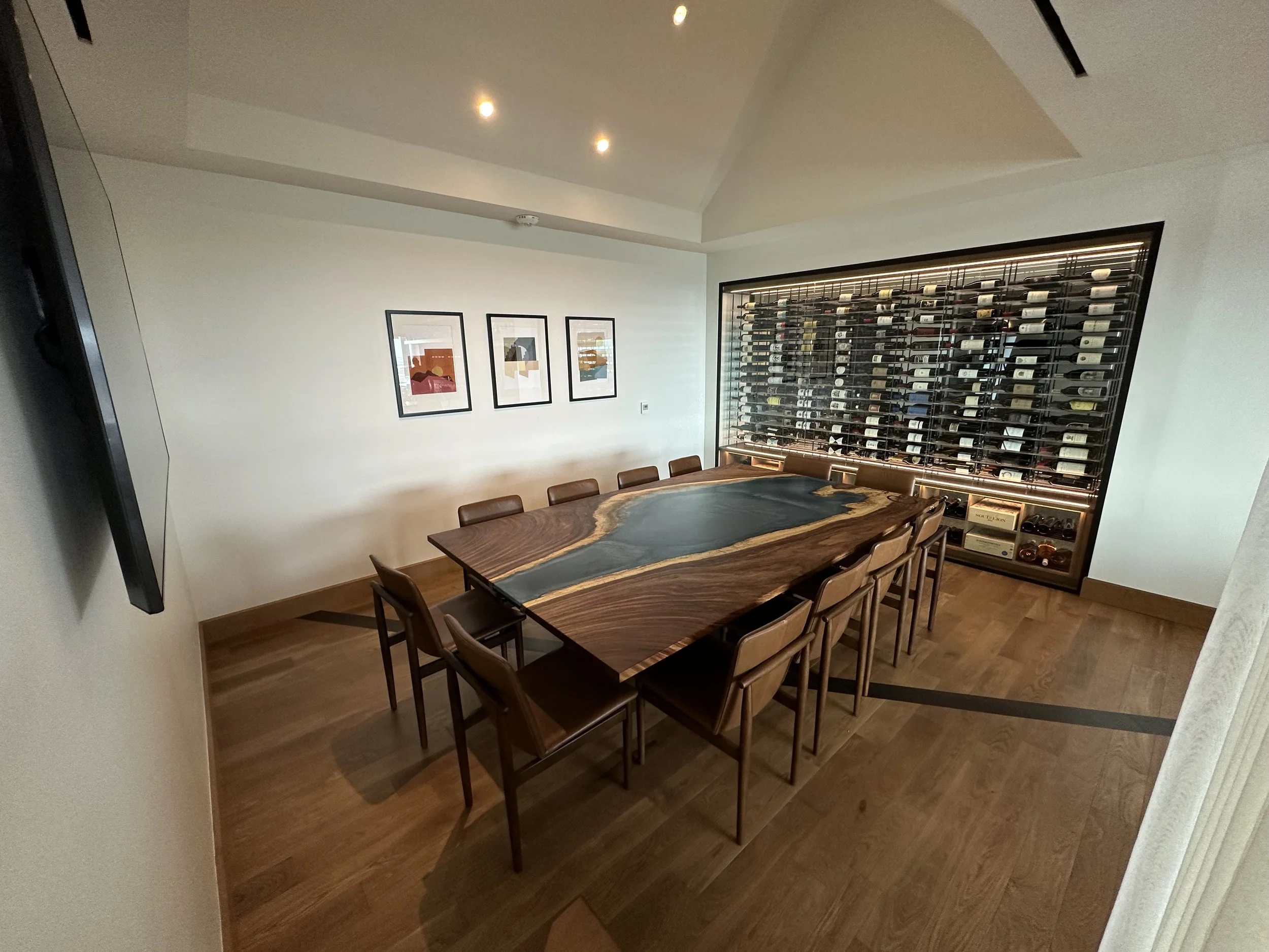Elegant dining room with a large wooden table featuring a river-like dark and tan pattern, surrounded by ten matching chairs. A wall-mounted wine rack filled with bottles covers most of the back wall. Three framed abstract art pieces hang on the left wall, and the room has recessed ceiling lights and hardwood floors.