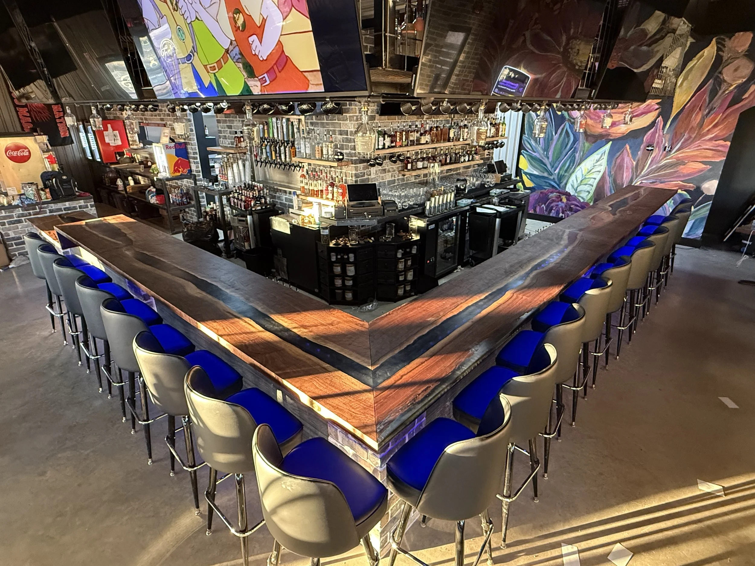 A modern bar with a large wooden U-shaped counter surrounded by blue and gray bar stools. The back wall features shelves with liquor bottles, and a colorful mural decorates one wall. There is a TV screen above the bar displaying animated characters.