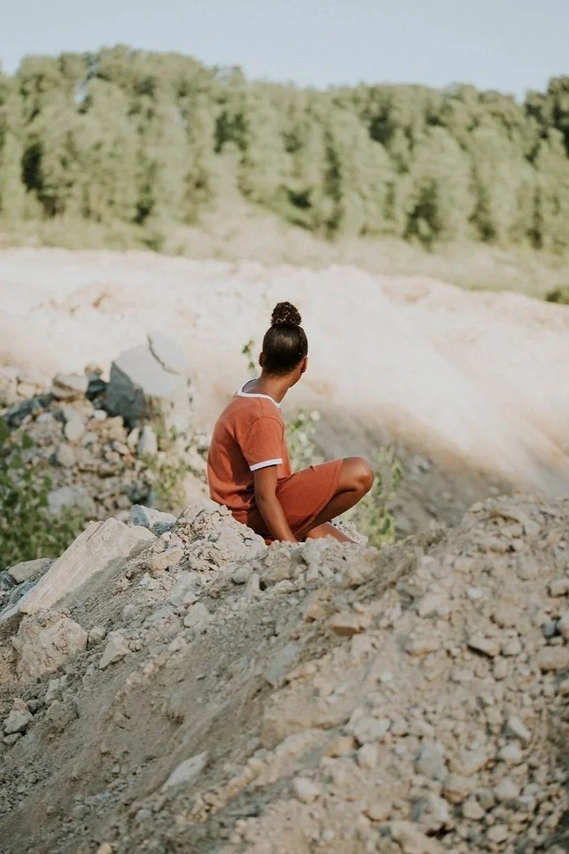 A young girl sits on a pile of dirt and rocks, looking out into a green, wooded landscape.