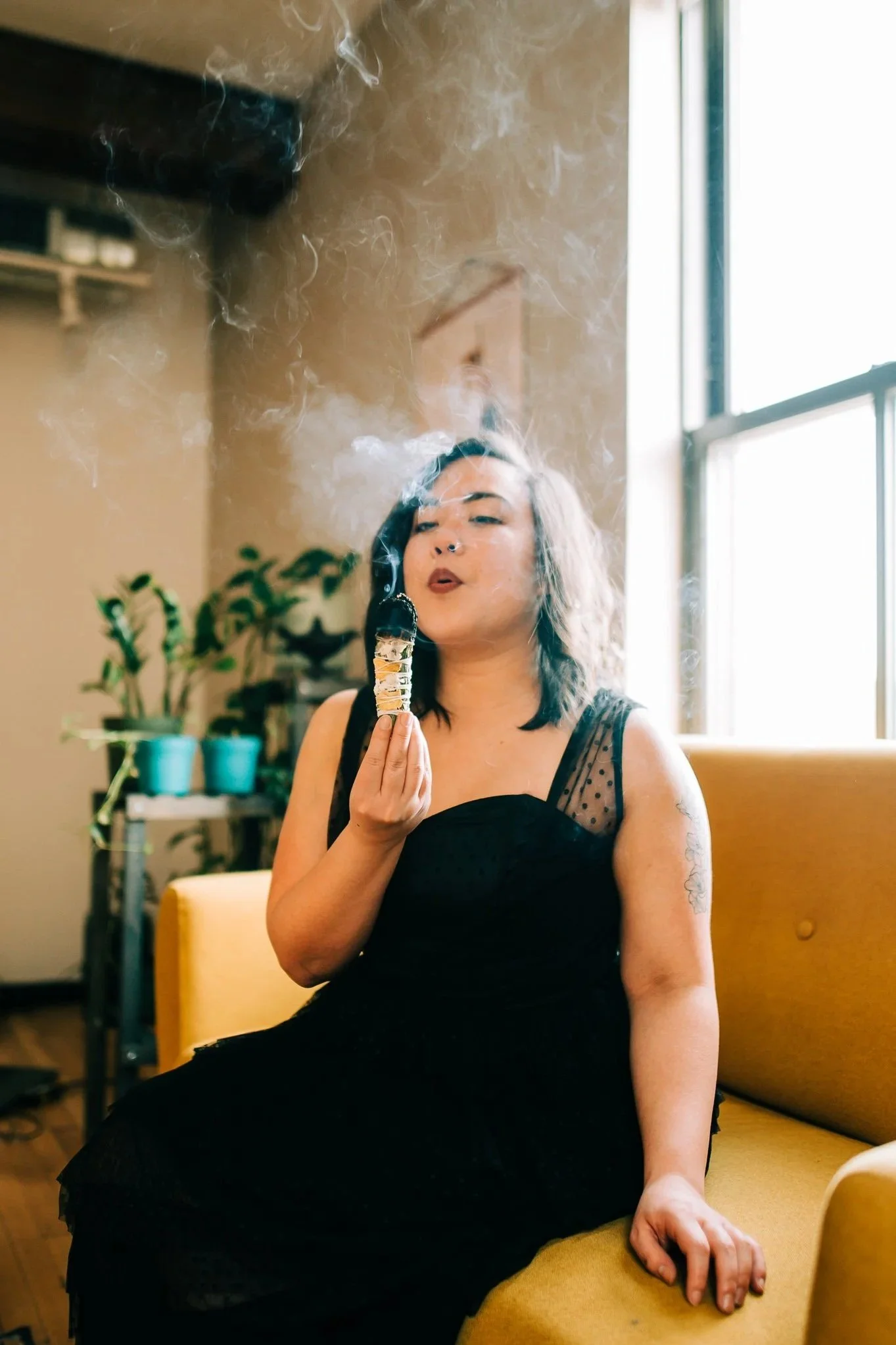 A woman in a black dress sitting on a mustard-colored couch, blowing out smoke from a cigar-shaped object, with a brightly lit window and houseplants behind her.