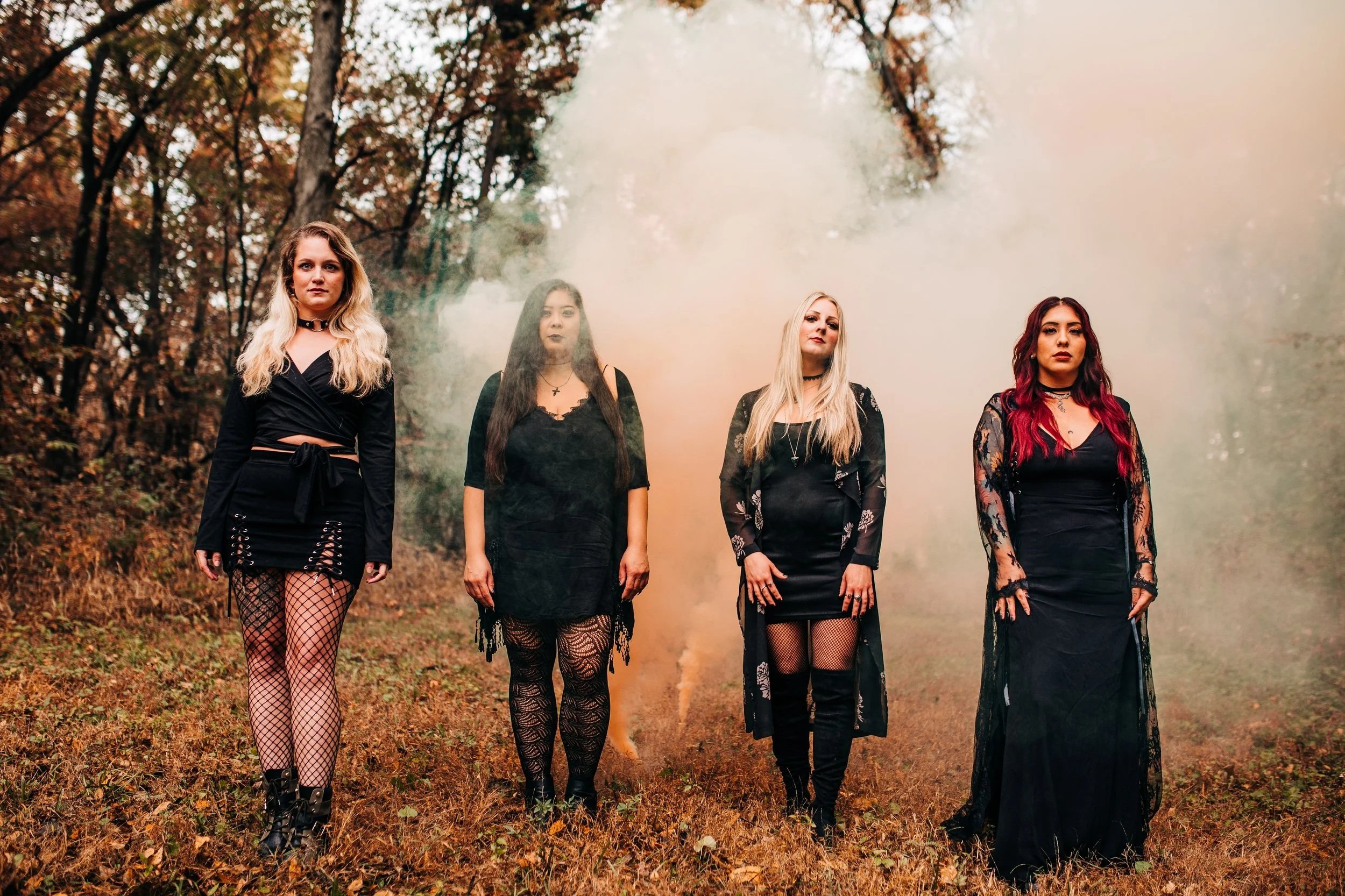 Four women dressed in black gothic or alternative fashion standing in a forest with autumn leaves, surrounded by smoke or fog.