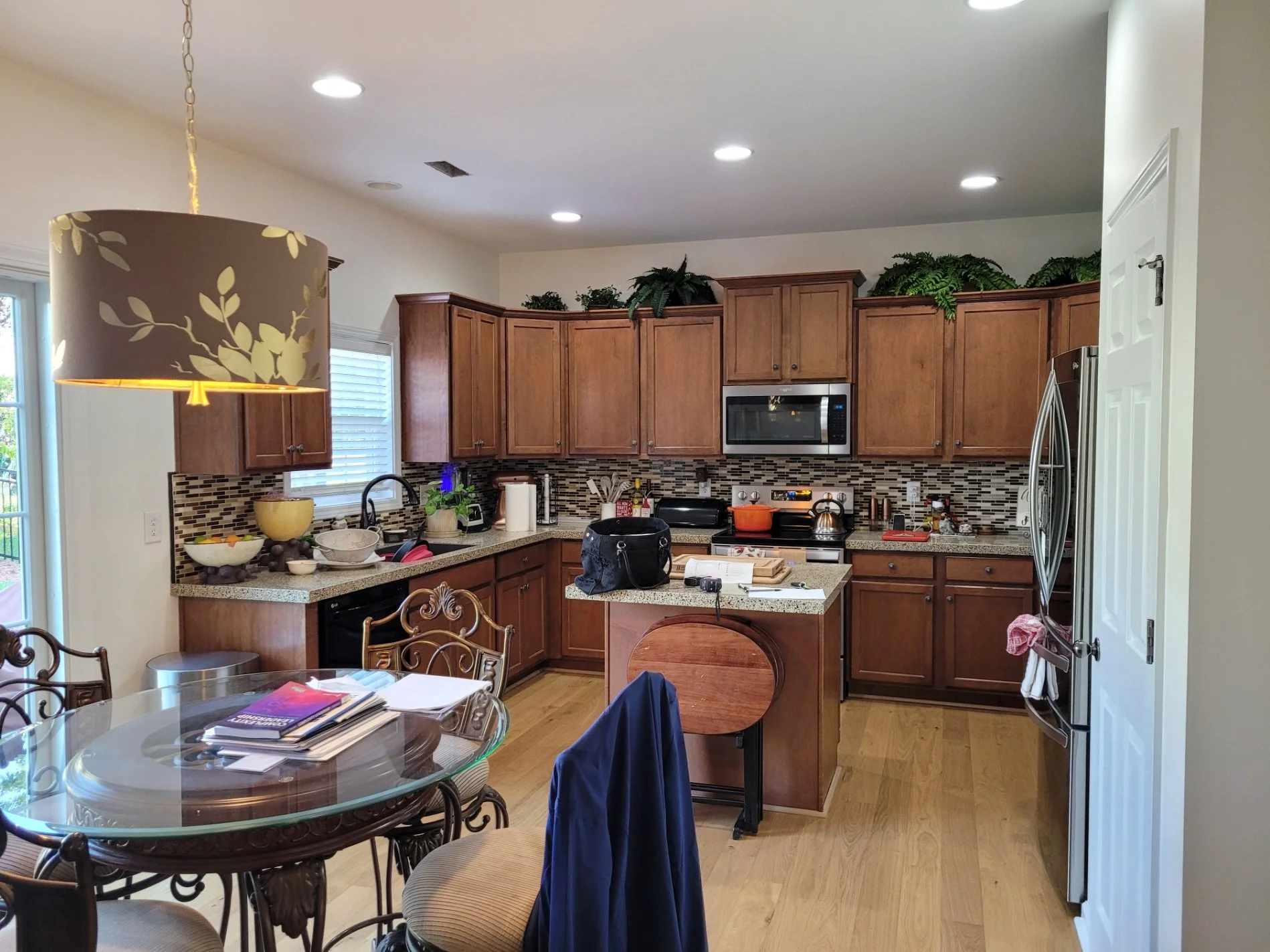 Kitchen with wooden cabinets, granite countertops, stainless steel appliances, and a mosaic tile backsplash. There is a small island with various items on it, a round glass dining table with four chairs, and green plants on top of the cabinets.