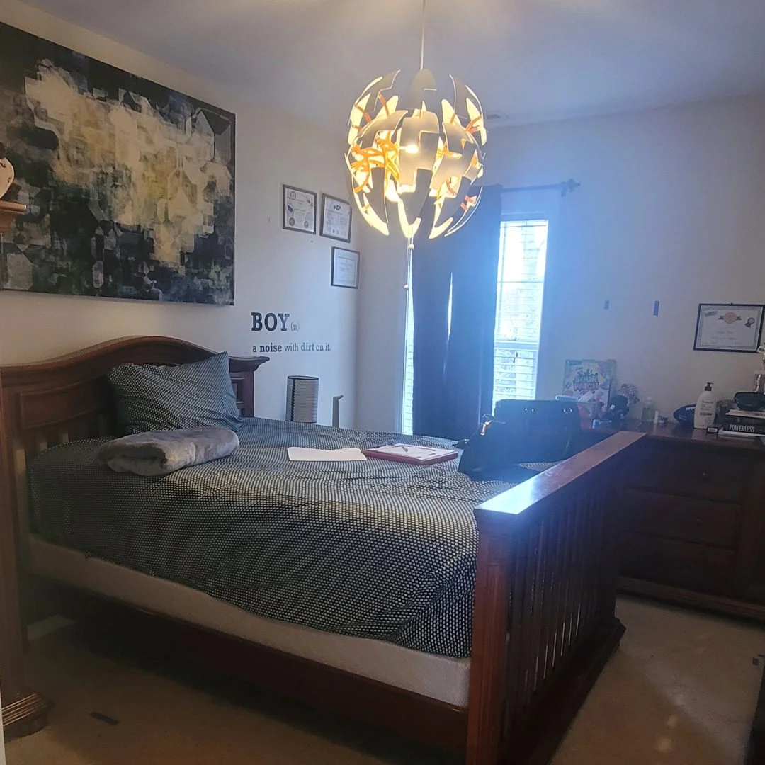 A woman in beige pants and coat standing in an empty, modern living room with light-colored wood flooring, a white marble fireplace, black framed windows, and a staircase with black railing.