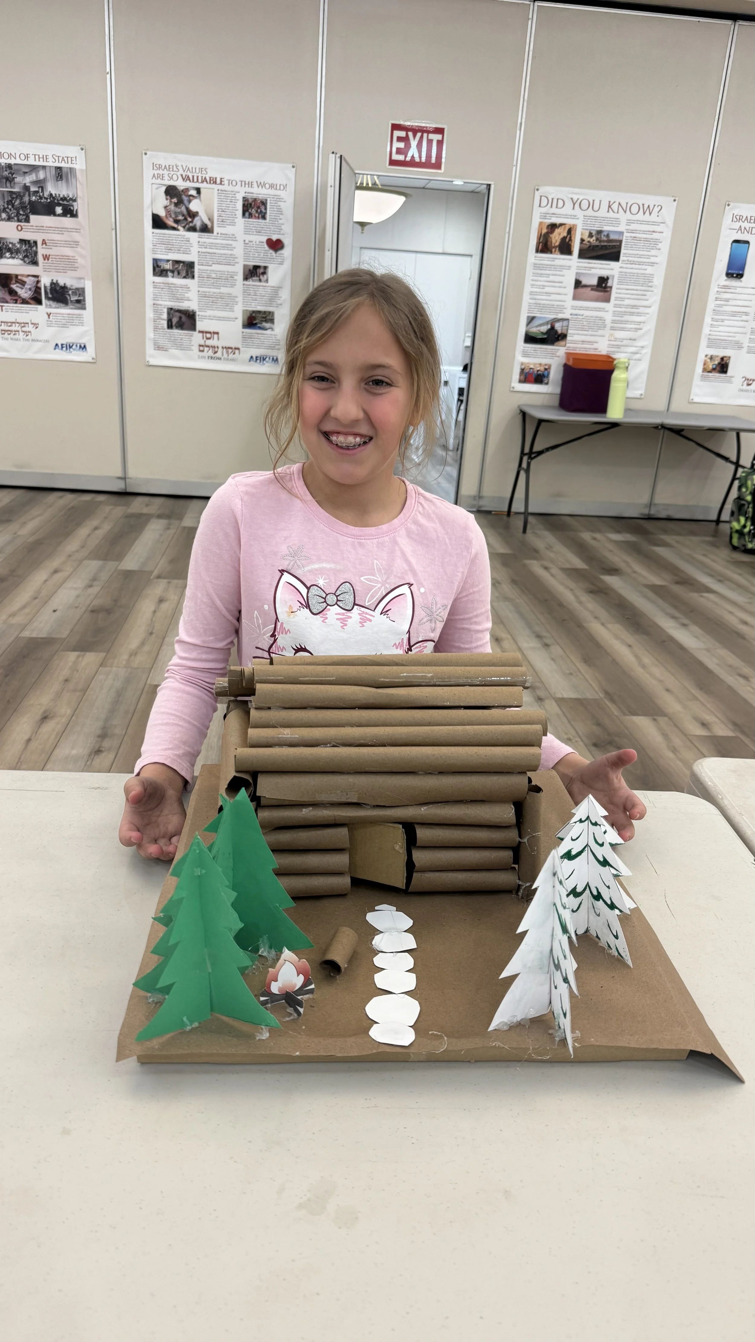 Smiling young girl with light brown hair in a pink long sleeve shirt standing behind a diorama of a log cabin in a forest scene. The diorama is made of cardboard and paper, featuring green and white paper trees and a pathway made of white paper circles, with a couple of small paper animals. The setting appears to be an indoor event with informational posters on the walls and an exit door in the background.