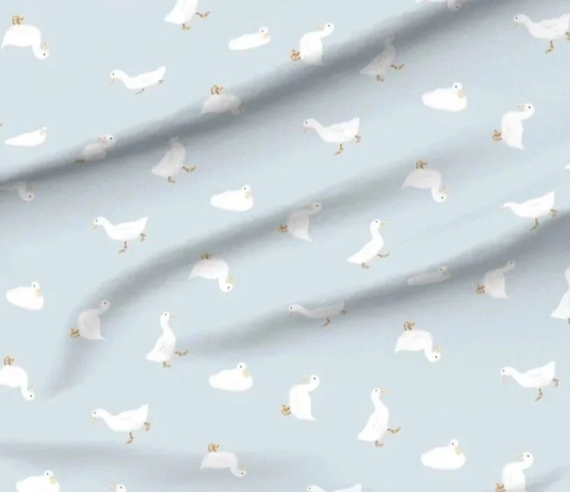 Playful whimsical fabric featuring scattered white ducks on a soft pastel blue background, shown as fabric with slight folds. The design has a light, airy feel suitable for nursery or kids’ rooms.