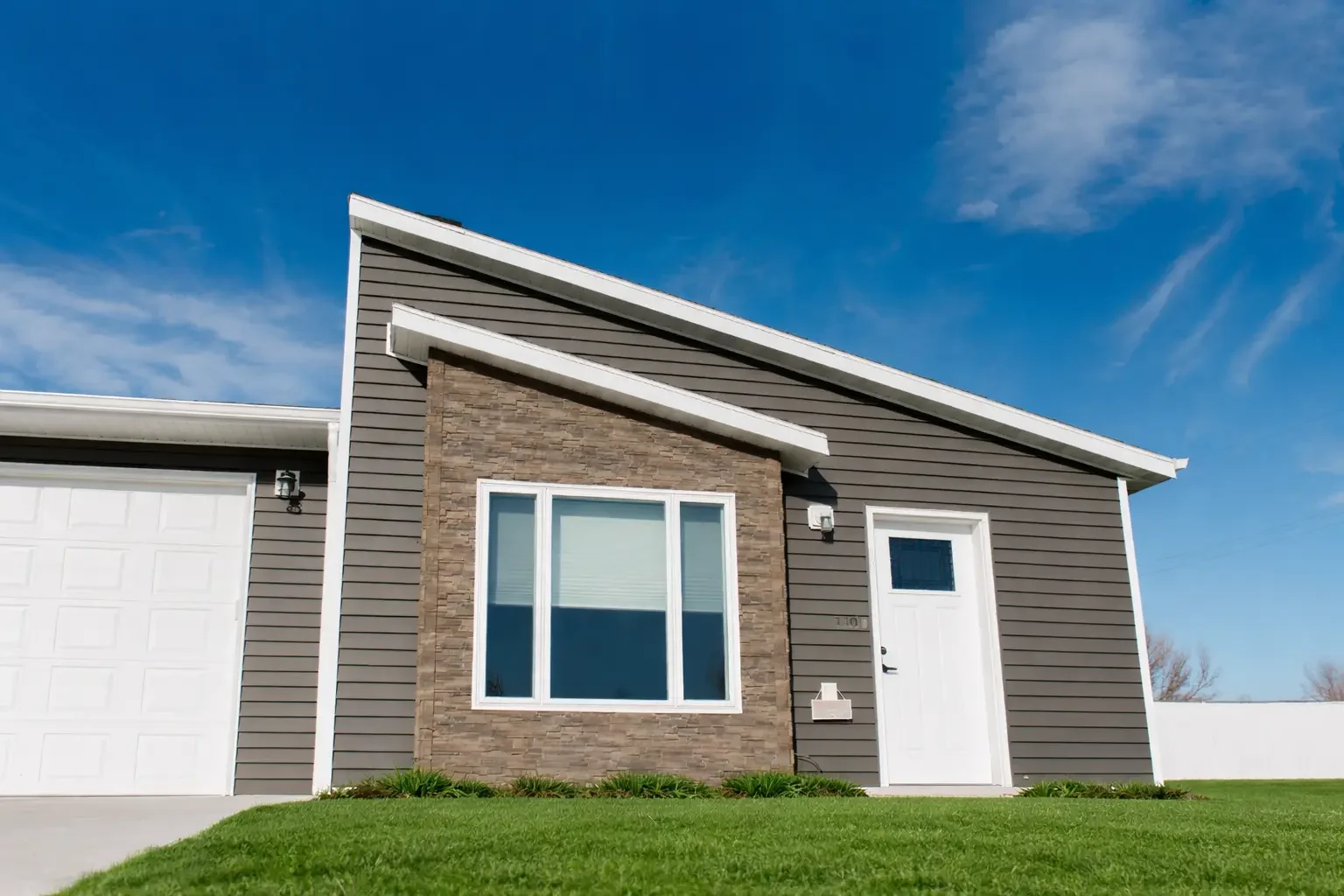Modern governor's house with a gray exterior, a white door, a large front window, a brick accent wall, and a well-maintained lawn under a blue sky.