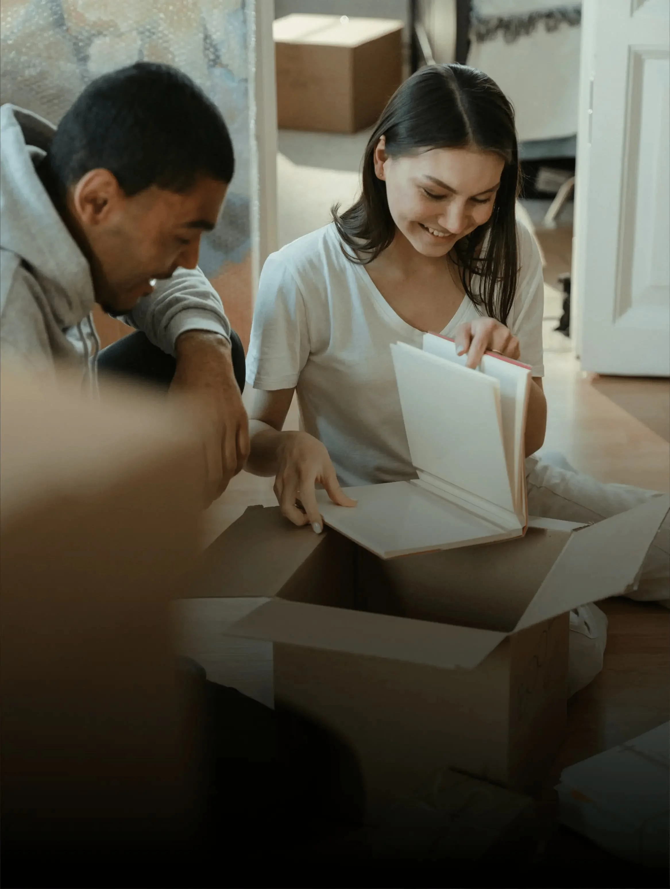 Two people sitting on the floor, smiling, looking at a book inside a cardboard box in a room with wooden flooring and household furniture in the background.